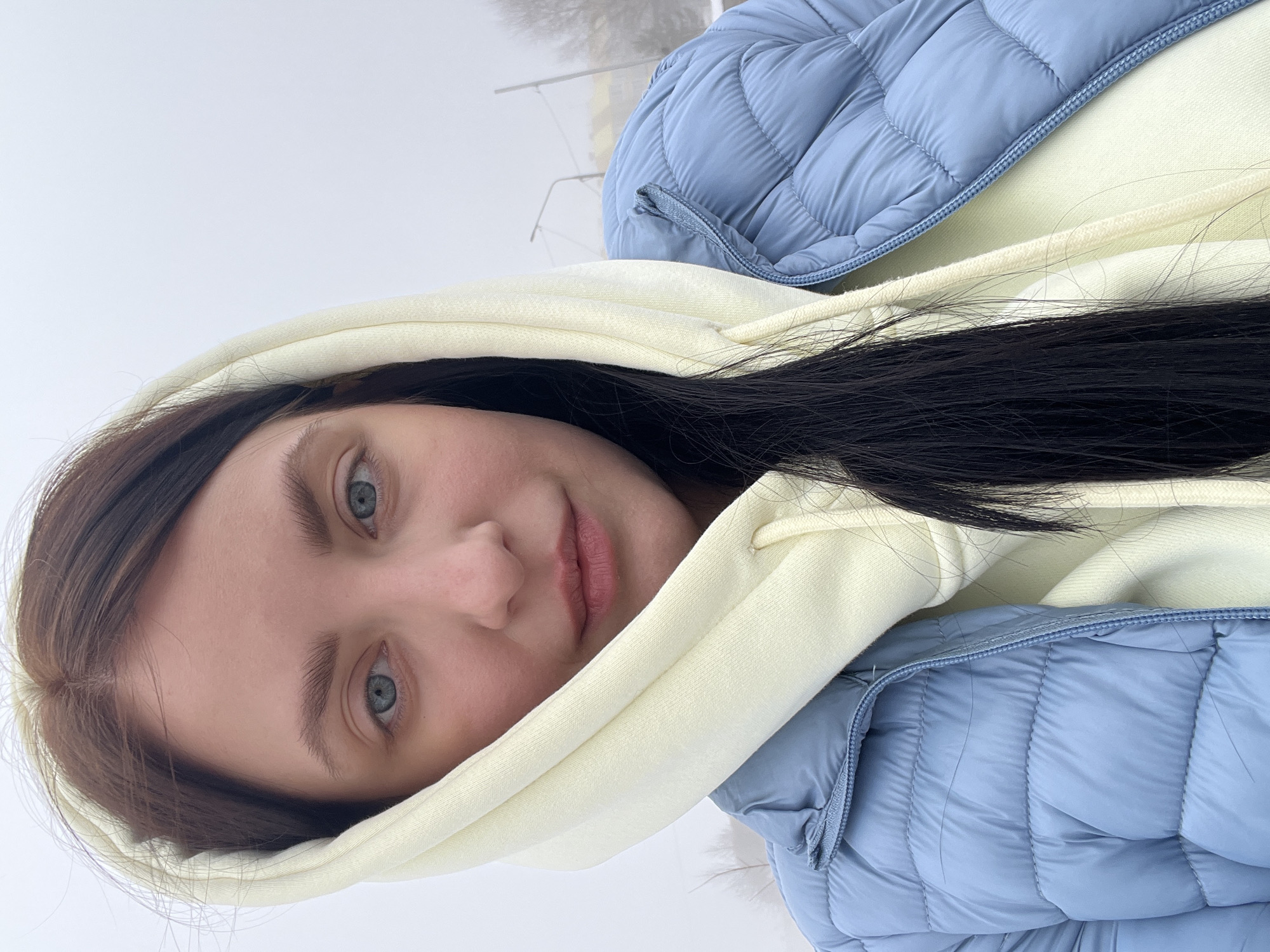 A young woman poses for a close-up outdoor portrait in what appears to be a foggy or overcast setting. She has fair skin, light blue eyes, and dark brown hair. She is wearing a light yellow hoodie with the hood up and a light blue quilted jacket. The location appears to be Марковский, Russia. In the background, indistinct structures or utility poles are visible, adding to the sense of a cold, possibly urban environment.