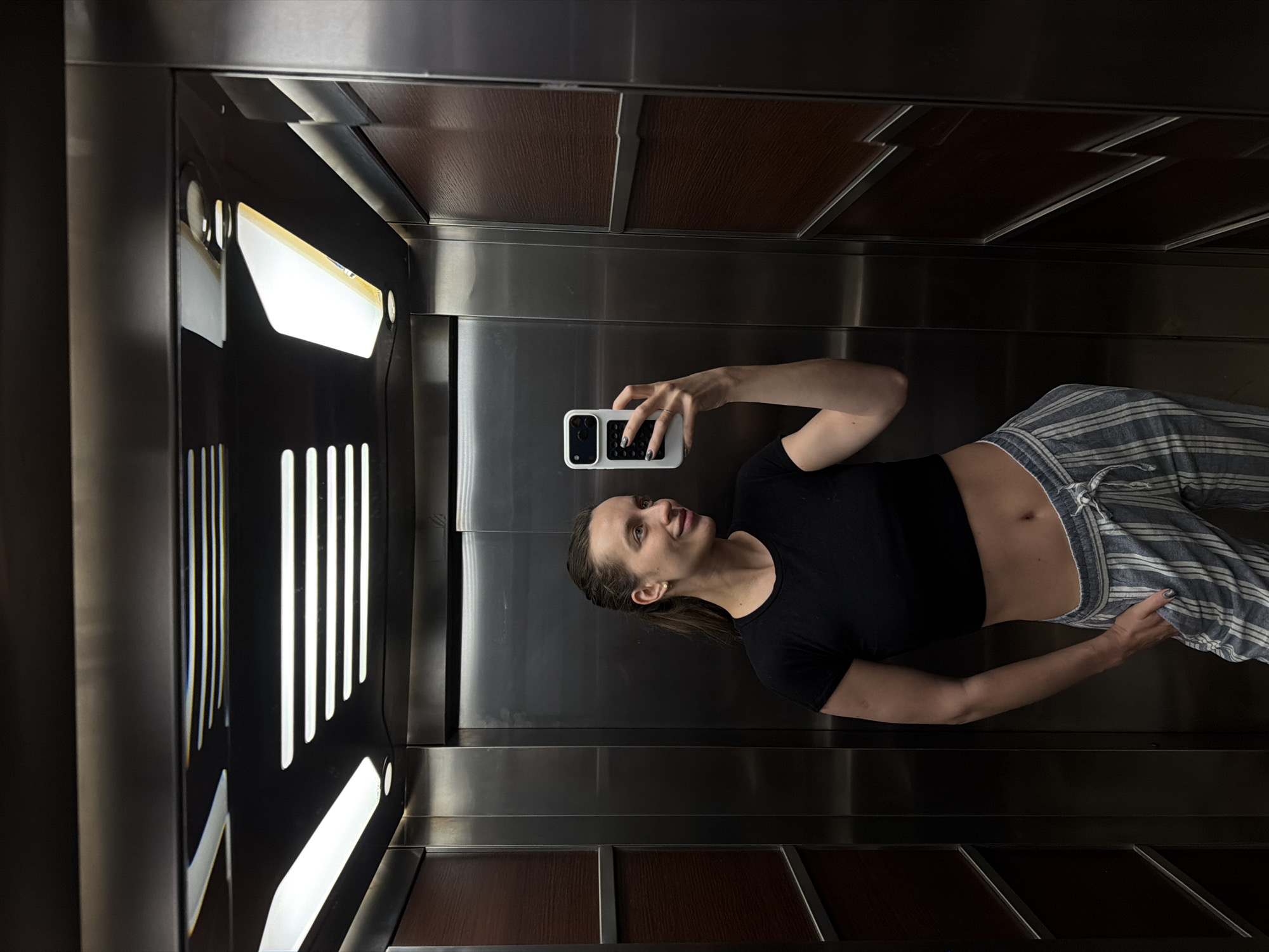 This media file captures a young woman taking a selfie inside a modern elevator, likely located in Budva, Montenegro. The image is presented in a rotated vertical orientation.
The subject is a woman with long, dark hair pulled back into a ponytail. She is wearing a short-sleeved black crop top that reveals her midriff, paired with loose-fitting blue and white striped drawstring pants. Her light-colored eyes are directed towards her smartphone, and a faint smile plays on her lips. She holds a white smartphone with a black camera module and distinct side buttons in her right hand, positioned to capture her reflection in the elevator's polished surfaces. Her fingernails appear to be painted a dark color.
The elevator interior features sleek, shiny stainless steel walls. Dark brown wooden panels with a visible grain line the ceiling and the lower section of the walls, adding a touch of warmth and contrast to the metallic surfaces. On the left side of the elevator, bright, vertical strip lights illuminate the space, casting reflections on the opposite wall, creating a sense of depth and modern design. The overall scene suggests a casual moment captured in a functional yet stylish environment. There is no visible text in the image.