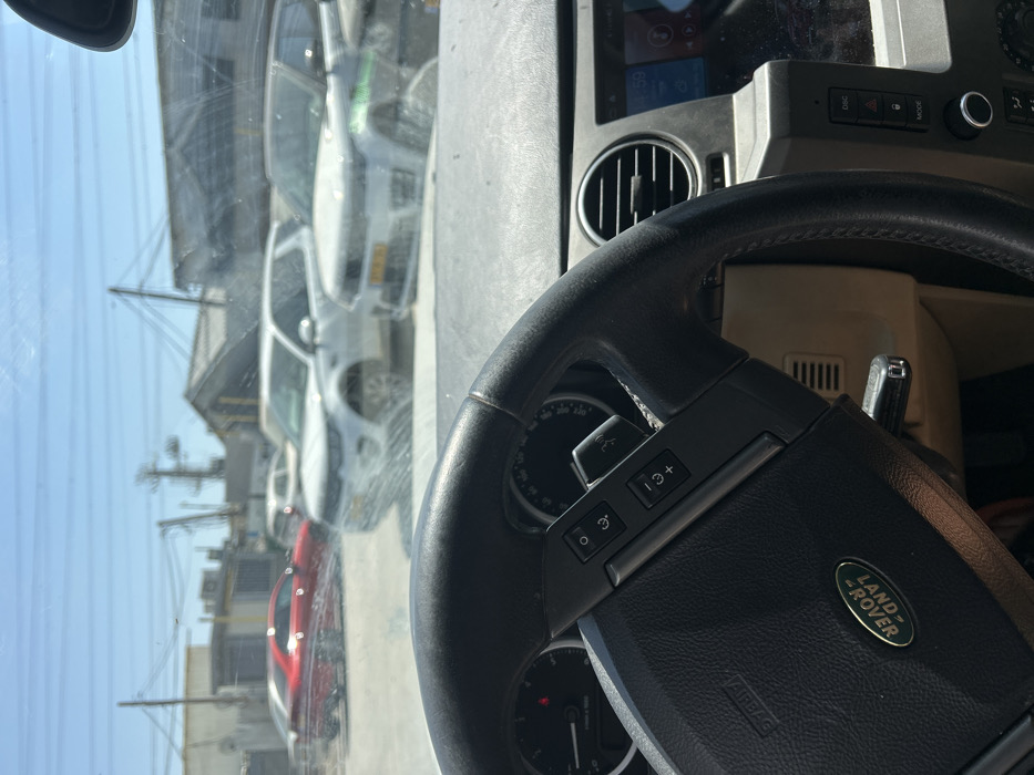 An interior shot of a Land Rover vehicle featuring the steering wheel prominently. The steering wheel has control buttons and a Land Rover logo. The dashboard with air vents and other controls is partially visible. Outside the vehicle, a row of cars, mostly white, is seen parked on a street with buildings and power lines in the background under a clear blue sky. The view is slightly obstructed by a dirty windshield.