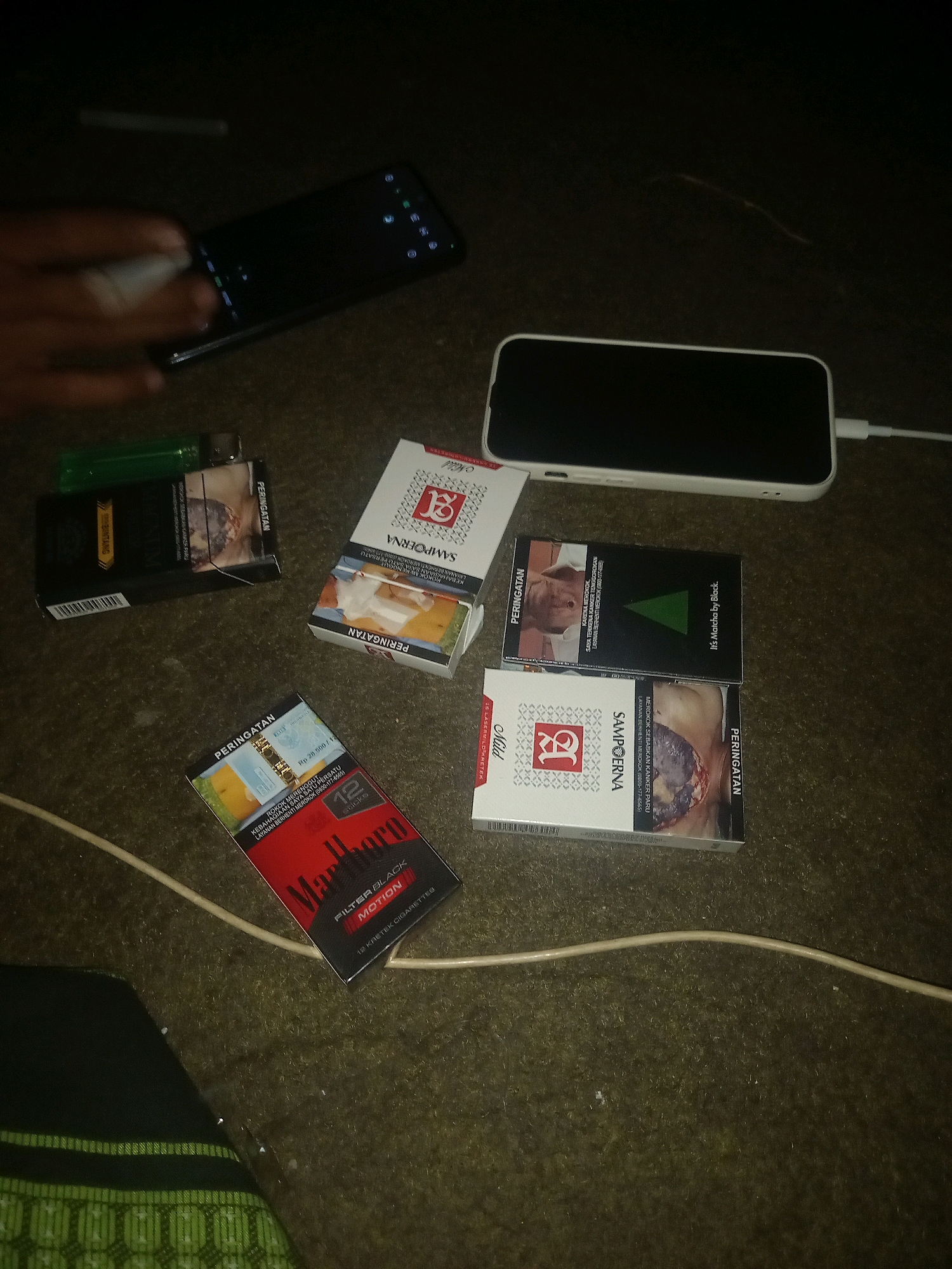 The image depicts a dimly lit, indoor scene, possibly a room, with a floor covered in a rough, textured material that appears to be a carpet or rug. Several objects are arranged on the floor, suggesting a casual or perhaps recreational setting.
Dominating the center of the frame is a white smartphone placed horizontally, with a white charging cable plugged into its right side, extending off-frame to the right. To the left of the smartphone, a black smartphone is visible, also lying flat. A hand, partially visible at the top left, appears to be interacting with the black smartphone, with a finger touching its screen. A light green lighter is also present to the left of the black smartphone, positioned next to a black cigarette pack.
Several cigarette packs are scattered around the scene. A red and black Marlboro "Filter Black Motion" cigarette pack is in the lower right quadrant, with a portion of a white cable running across it. To the left of the Marlboro pack, a white cigarette pack with red and gold accents, labeled "Sampoerna Mild," is visible. Another cigarette pack, also white with red and gold accents and labeled "Sampoerna," is positioned to the upper right of the Marlboro pack. A black cigarette pack with a green triangular logo and text that reads "It's Mild by Black." is situated to the right of the white Sampoerna pack.
The lighting in the image is low, with the primary light sources likely originating from the smartphones and potentially an overhead light not fully captured. The overall impression is one of a person or persons engaged with their mobile devices, with cigarettes readily available. The presence of the cigarettes and the setting do not provide specific clues to a precise location within Paiton, Indonesia, other than it being an indoor environment.