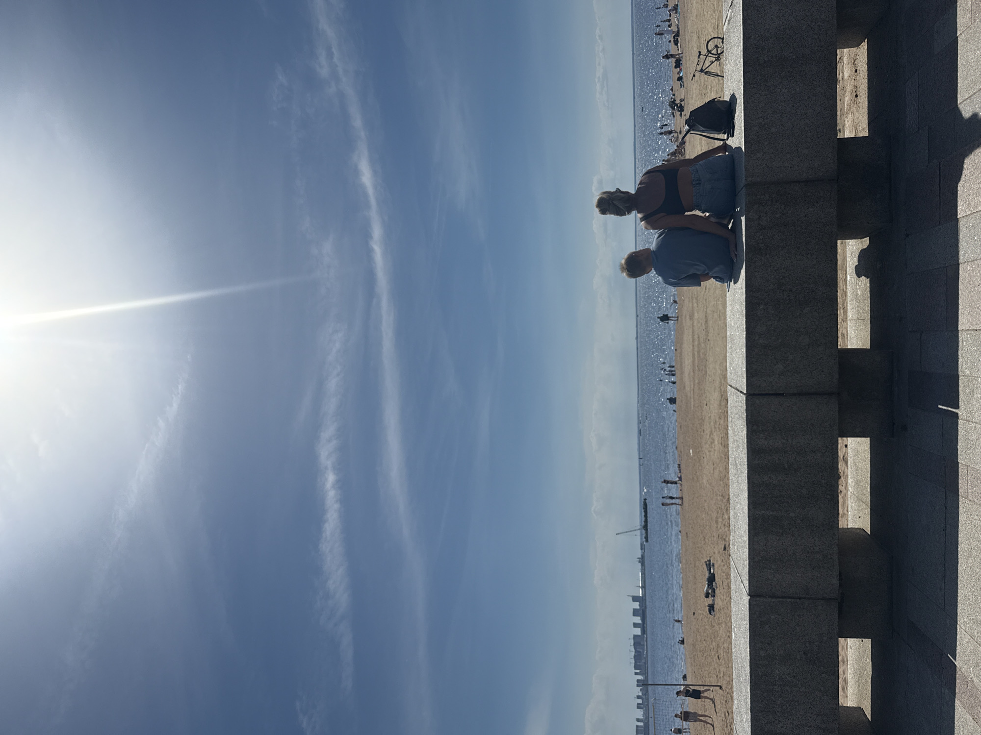 The image captures a sunny day at a beach in Saint Petersburg, Russia, with a clear blue sky and a bright sun. In the foreground, a woman and a young boy are seated on a concrete wall, facing the water and the distant cityscape. The woman wears a black top and denim shorts, with her hair tied up. The boy is dressed in a blue t-shirt and shorts. A black backpack rests beside them on the wall, and a bicycle is parked to the right.
The beach in the midground is populated with various individuals, some walking, others sitting or lying on the sand. A volleyball net is visible on the left side of the beach. The water is calm, with a few small boats scattered across it. In the background, the Saint Petersburg skyline is visible, characterized by tall buildings and construction cranes, suggesting ongoing development. The scene conveys a relaxed atmosphere of a summer day at the beach.