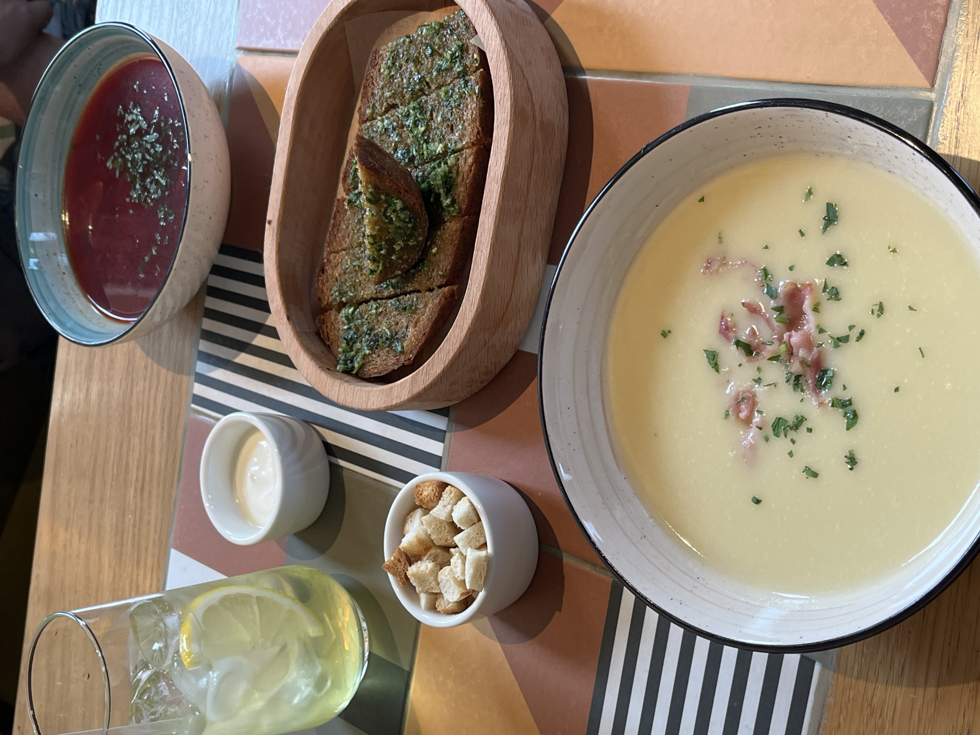 A top-down shot displays a variety of food items on a multicolored table, which includes stripes and squares of white, black, orange, and brown, in an establishment in Verkhnyaya Pyshma, Russia. A glass of lemonade with a lemon wedge and ice is at the bottom left. To the right of the drink is a small bowl of white sauce and a bowl of croutons. Moving upwards, the image shows a bowl of red soup, a wooden bowl filled with slices of garlic bread topped with herbs, and a bowl of creamy white soup garnished with green herbs and ham pieces. The food is arranged in a line from left to right on the table.