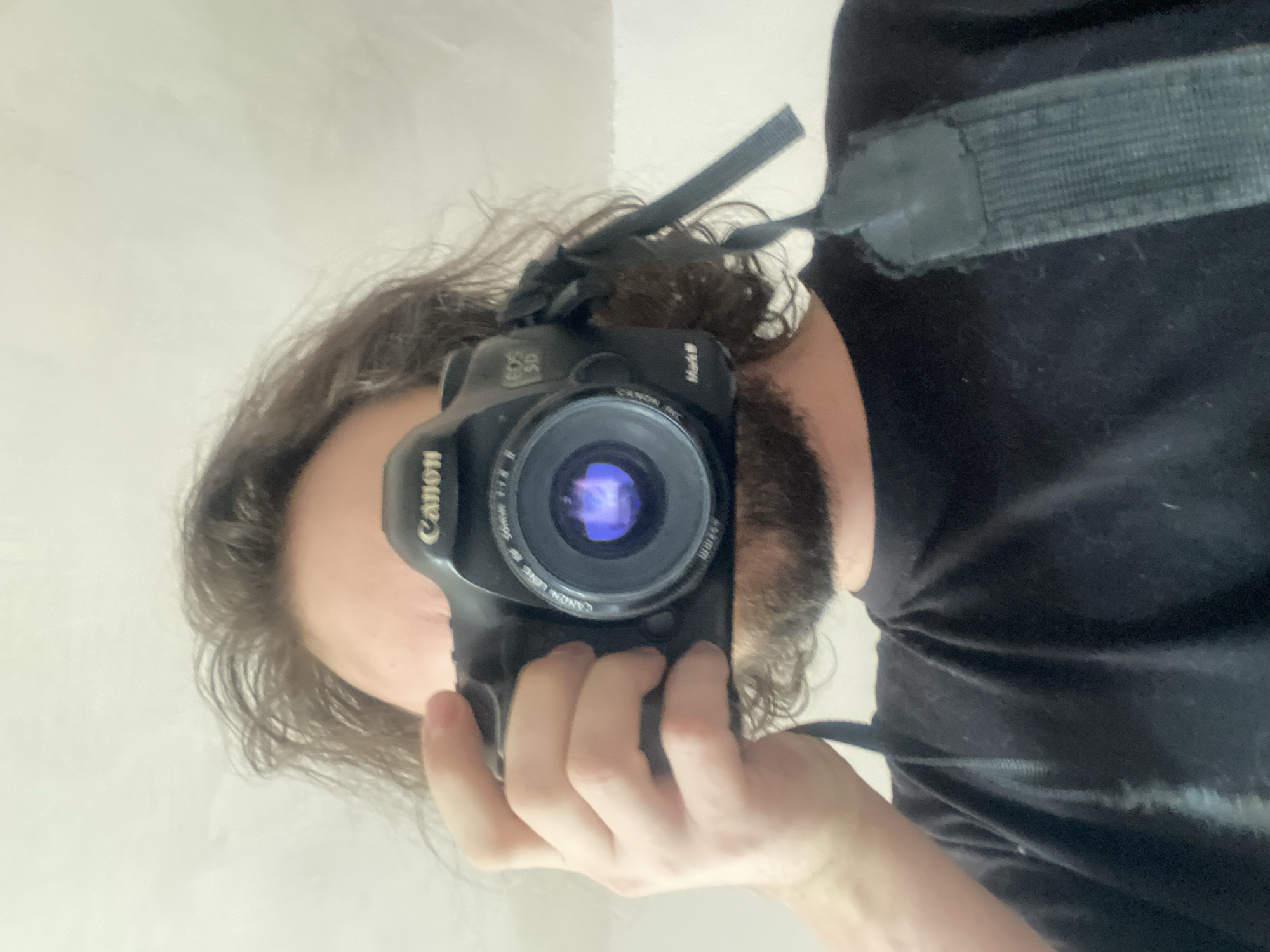 A man is taking a selfie with a black Canon DSLR camera. He is wearing a black shirt and the camera strap is visible. Part of his long, curly hair and beard is visible. The background is a plain, light-colored wall.
