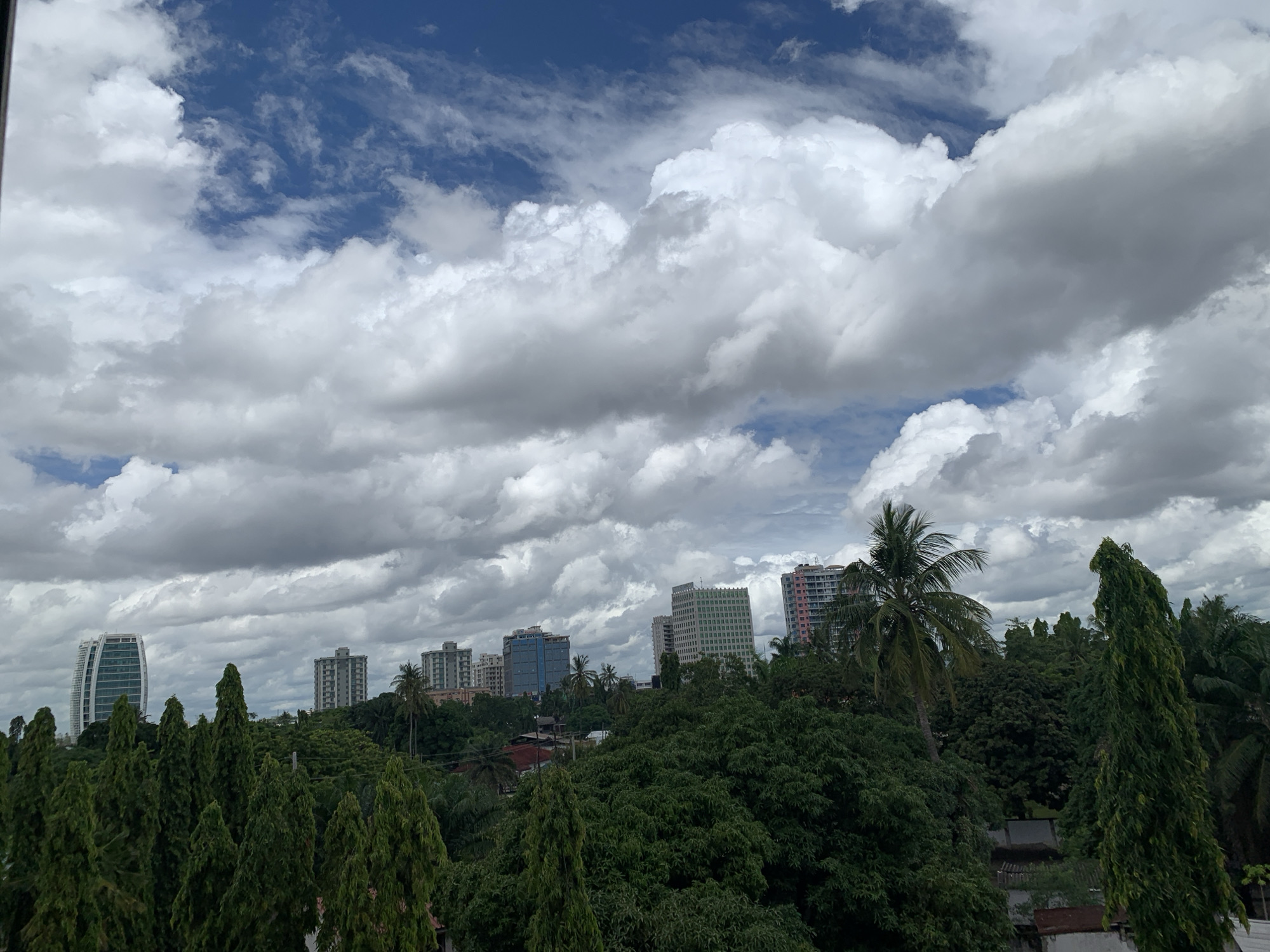 The image captures a cityscape under a sky filled with scattered cumulus clouds, suggesting a partly cloudy day. The visible architecture includes several high-rise buildings and residential structures. A dense collection of trees, including palm trees, occupies the foreground and middle ground, obscuring some of the buildings and residential houses behind them. The location is most likely Dar es-Salaam, Tanzania, evidenced by the architectural styles and tropical vegetation characteristic of the region. There are no individuals visible in the image.