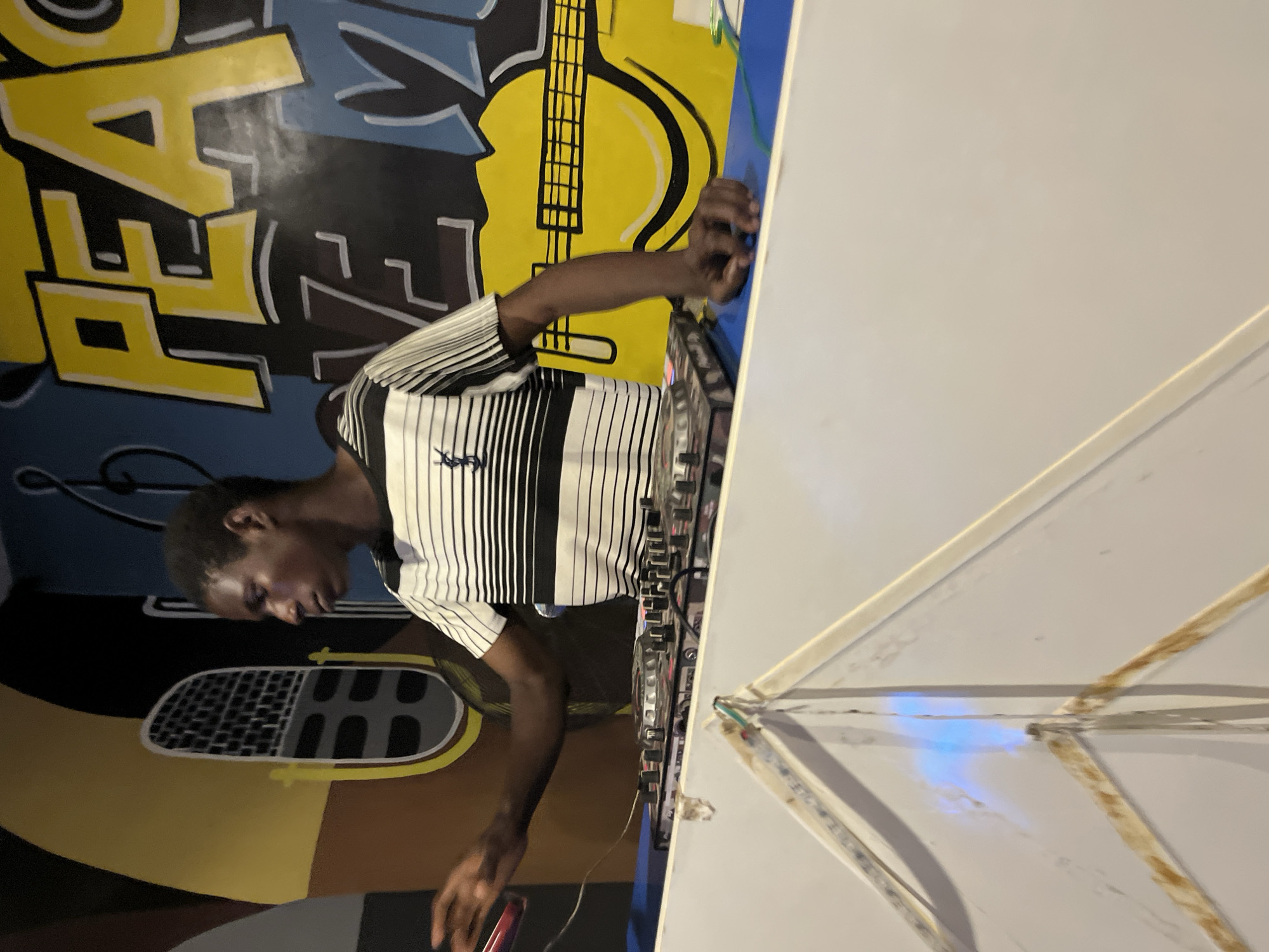 A young Black male is standing behind a DJ booth, facing to the right, with his right arm extended as if playing music. He is wearing a black and white striped shirt. In front of him is a DJ mixer with various knobs and buttons. The background features a brightly colored mural with a stylized microphone, the word "PEACE" in large yellow letters, and a yellow guitar. The environment appears to be an indoor venue, possibly a club or performance space, with blue walls visible behind the mural.