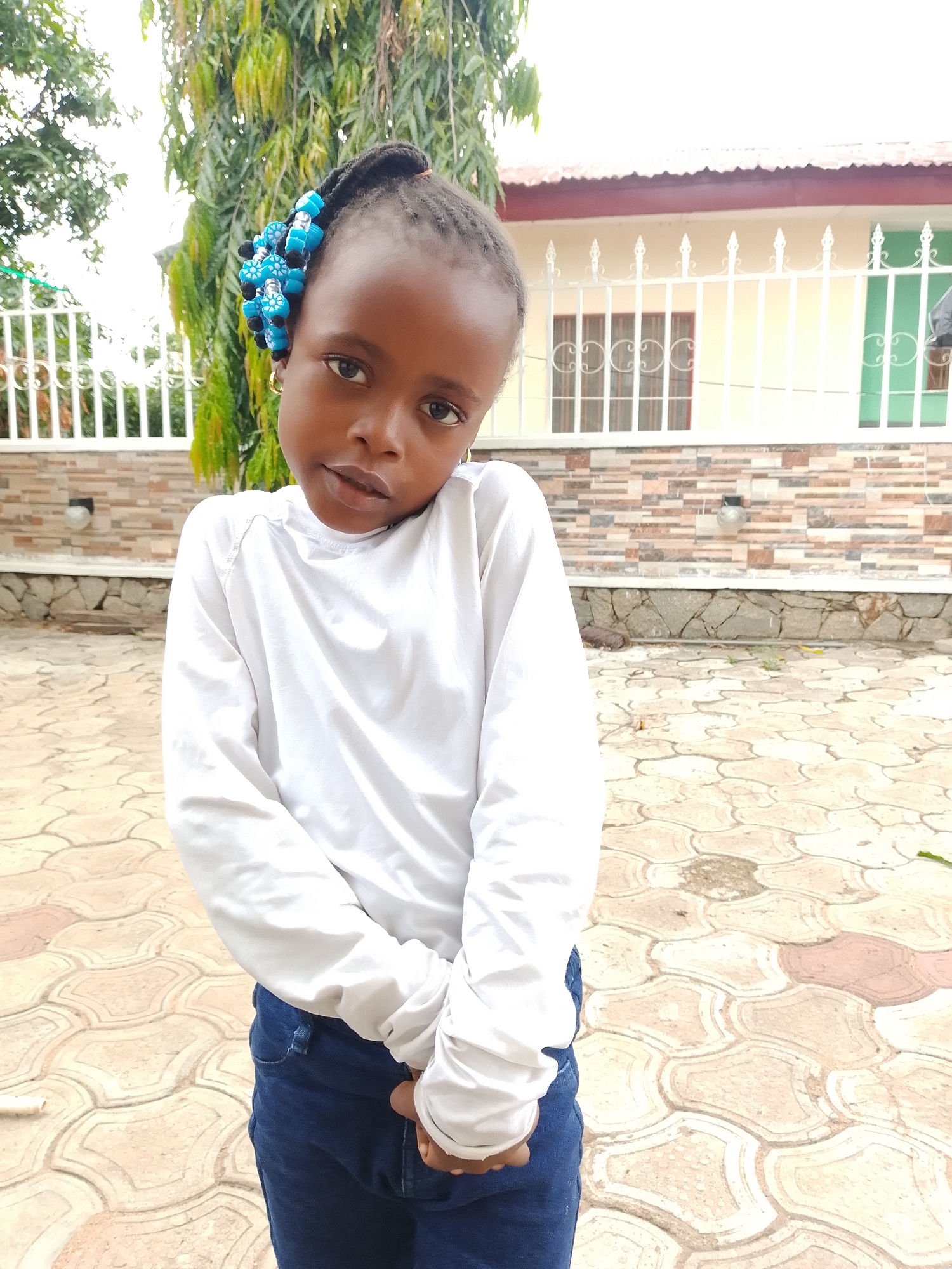 A young Black girl stands centrally in an outdoor, daylight portrait. She has dark skin and her hair is styled in braids, with a prominent cluster of blue and black beaded decorations on the right side of her head. Small gold hoop earrings are visible in her ears. She is wearing a plain white long-sleeved shirt and blue jeans, with her hands clasped together in front of her. Her head is tilted slightly to her right, and she looks directly at the camera with a neutral to slightly serious expression.
The setting appears to be a paved courtyard or yard. The ground is covered with light-colored, interlocking paving tiles in various shades of beige and light brown. Behind the girl, there's a low stone wall leading to a decorative white metal fence. Beyond the fence, a house is visible with a light-colored (possibly cream or beige) facade, a reddish-brown roof, and several windows with decorative metal grilles. The lower portion of the house's wall has a textured, stone-like finish. Lush green foliage and trees are prominent in the background, particularly on the left side, suggesting an abundance of vegetation. The lighting is bright and even, indicating daytime. There is no visible text in the image.