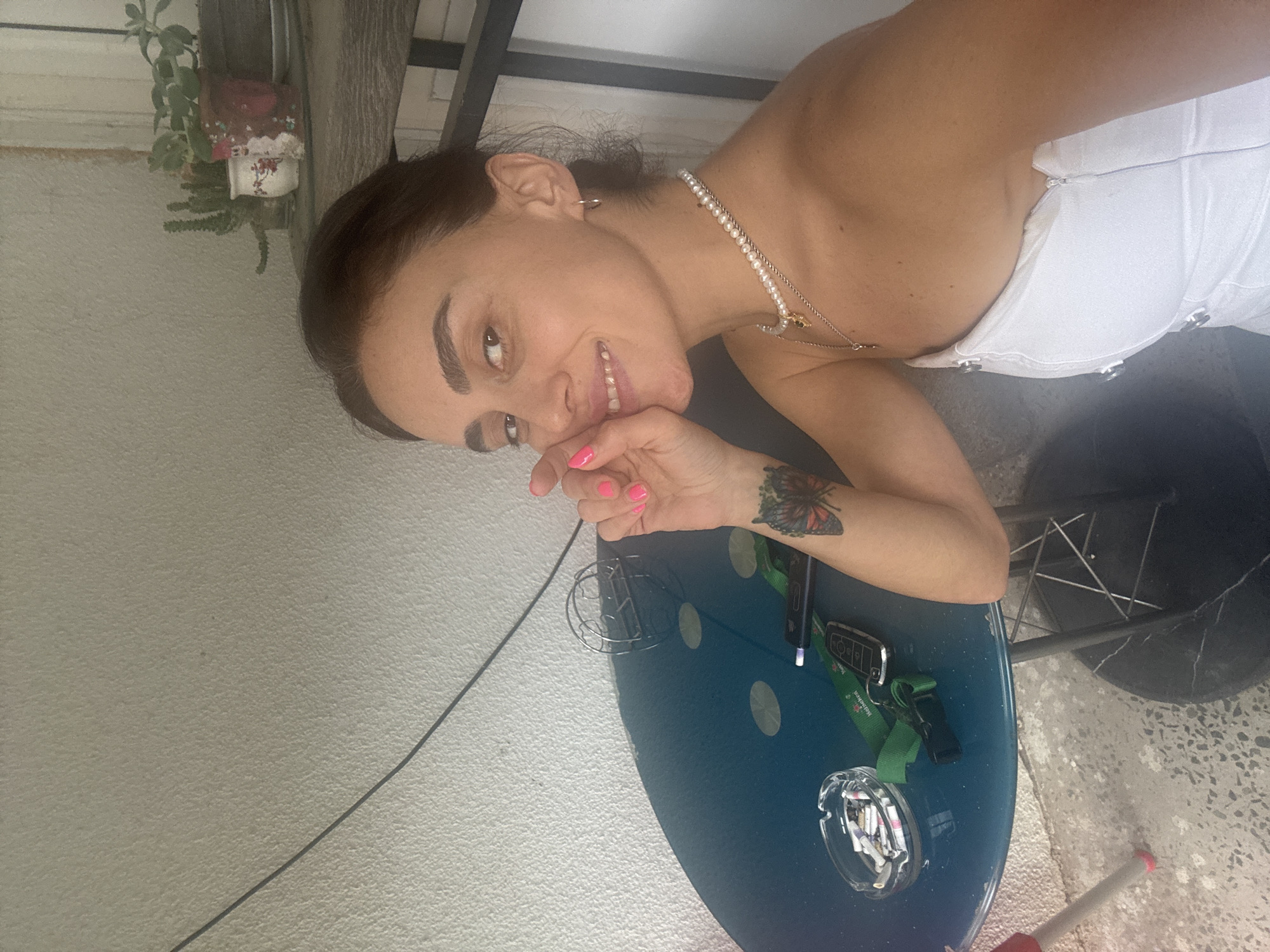 A woman with dark hair and light skin is pictured sitting at a blue round table. She is wearing a white strapless top and a pearl necklace. Her right arm is resting on the table, and she has a tattoo of two butterflies on her forearm. On the table are an ashtray filled with cigarette butts, a car key, a green lanyard with a Heineken logo, and a black electronic device. The woman is smiling and touching her face with her right hand, which has bright pink nail polish. In the background, there is a textured white wall and a shelf with several potted plants, including cacti. The scene appears to be outdoors on a balcony or patio, possibly in Tbilisi, Georgia.