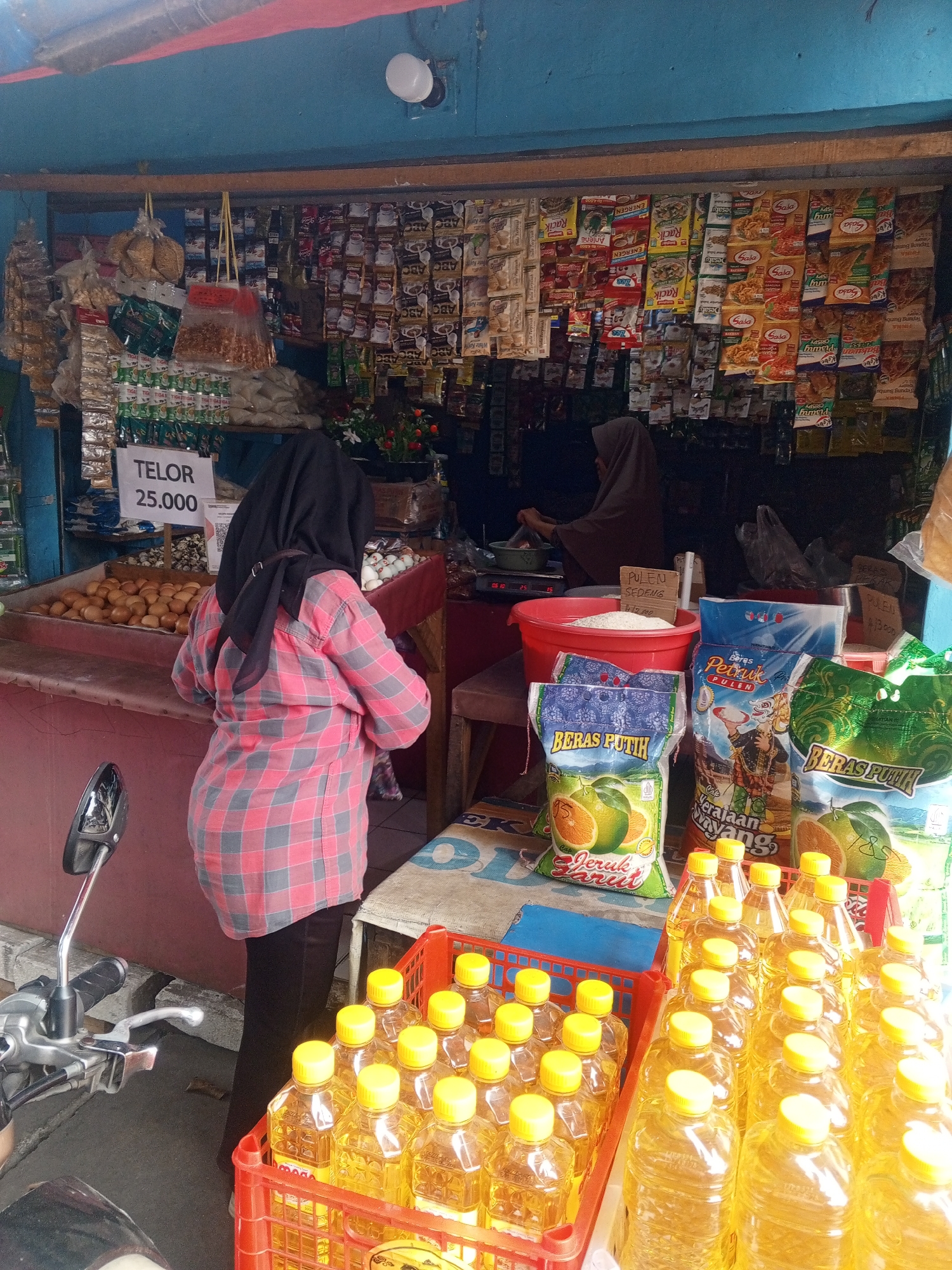 In Depok, Indonesia, a street-side shop displays a variety of goods. A person wearing a pink and grey checkered shirt and black pants, with a black hijab, stands facing a stall filled with eggs. A sign above the eggs reads "TELOR 25.000". To the right of the eggs, another person, wearing a beige hijab, is seen behind the counter, engaged in activity. Shelves behind them are stocked with numerous packaged food items, including instant noodles and spices. In the foreground, a red crate overflows with bottles of cooking oil with yellow caps. A motorcycle is partially visible on the left side of the image. The scene is illuminated by natural light, suggesting it is daytime.