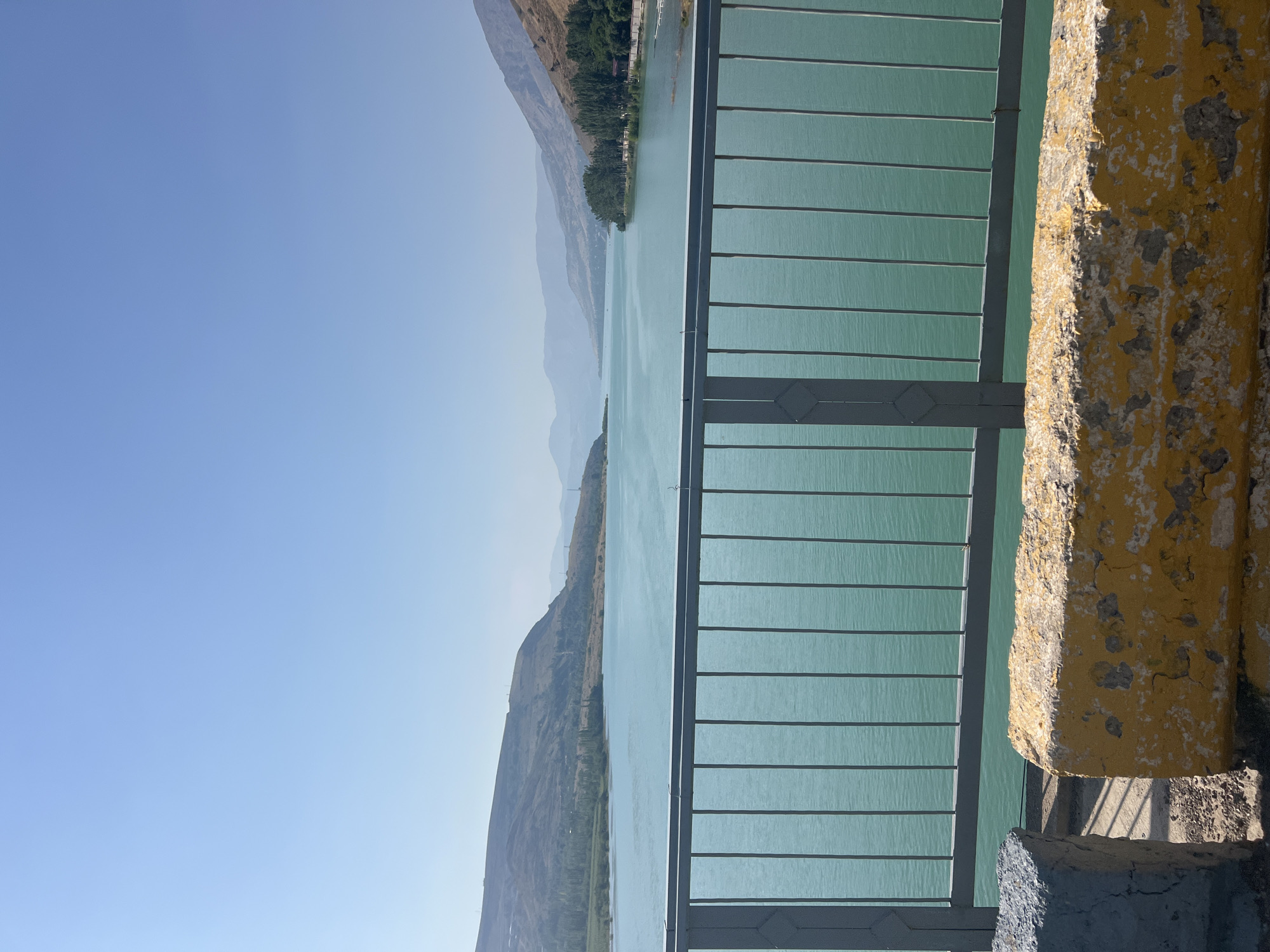 The image depicts a scenic view of a large body of turquoise water, likely a reservoir or lake, surrounded by rolling, arid hills. In the foreground, a grey metal railing runs horizontally across the frame, obscuring the lower portion of the water. Beyond the railing, two weathered concrete barriers are visible on the left side of the frame, one grey and the other painted yellow and white, showing signs of age and wear. The water's surface is calm, with subtle ripples reflecting the clear blue sky above. Distant hills and mountains, some sparsely covered with vegetation, frame the expansive water. On the right side of the water, a line of trees suggests a developed area, possibly a park or settlement. The overall impression is one of a tranquil, natural landscape under a bright, clear sky. Based on the prompt, this scene is located in the city of Yanghikurgan, Uzbekistan.