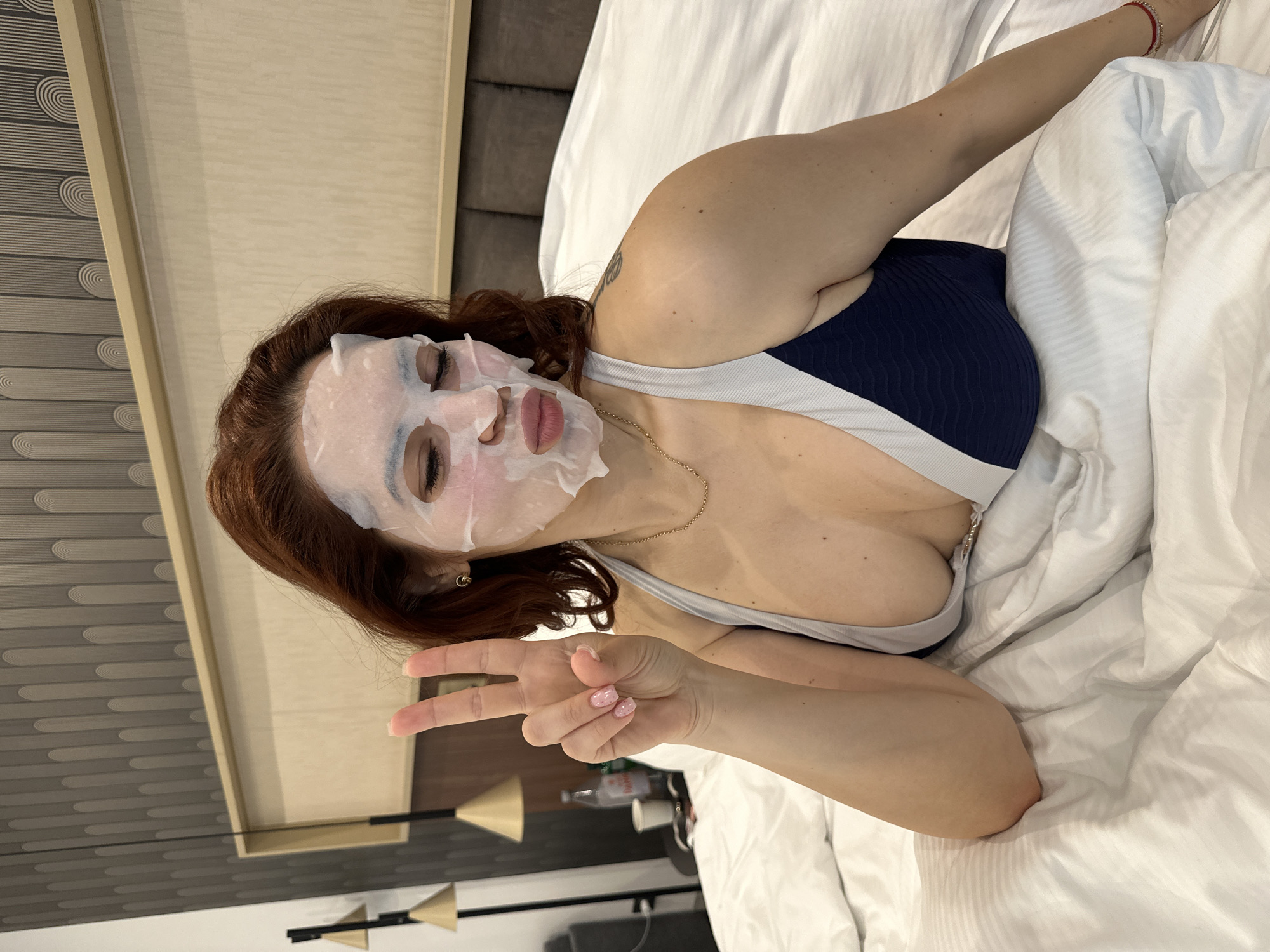 A woman with reddish-brown hair and fair skin is lying in a bed covered with white sheets. She has a white face mask applied and is wearing a navy blue and white sports bra. She is making a peace sign with her left hand. A gold necklace adorns her neck. A bedside table with a lamp, water bottle, and cup is visible in the background. The scene does not include any information to confirm it is in Kazanlak, Bulgaria.