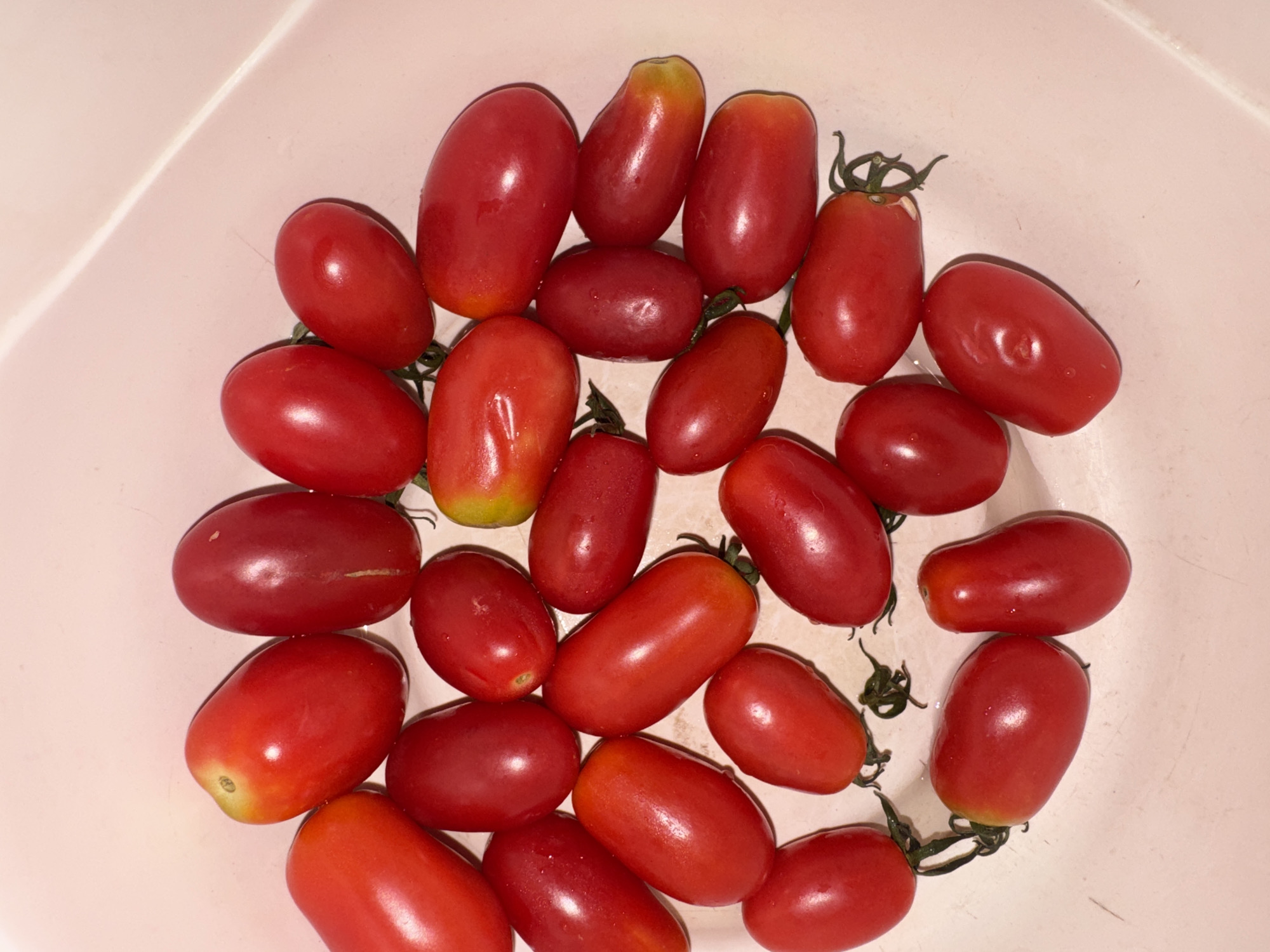 A top-down, medium shot captures a cluster of ripe, red cherry tomatoes, some with slight yellowing near the stem, nestled in a light pink bowl. The tomatoes are oval and plump, with a glossy sheen indicating freshness. A few small green stems and calyxes are visible, attached to some of the tomatoes. The background is a plain, light pink surface, likely the interior of the bowl, with a subtle diagonal line suggesting the bowl's rim or a crease. The lighting is somewhat harsh, creating bright highlights on the tomatoes. No people or specific city locations are identifiable in this image.