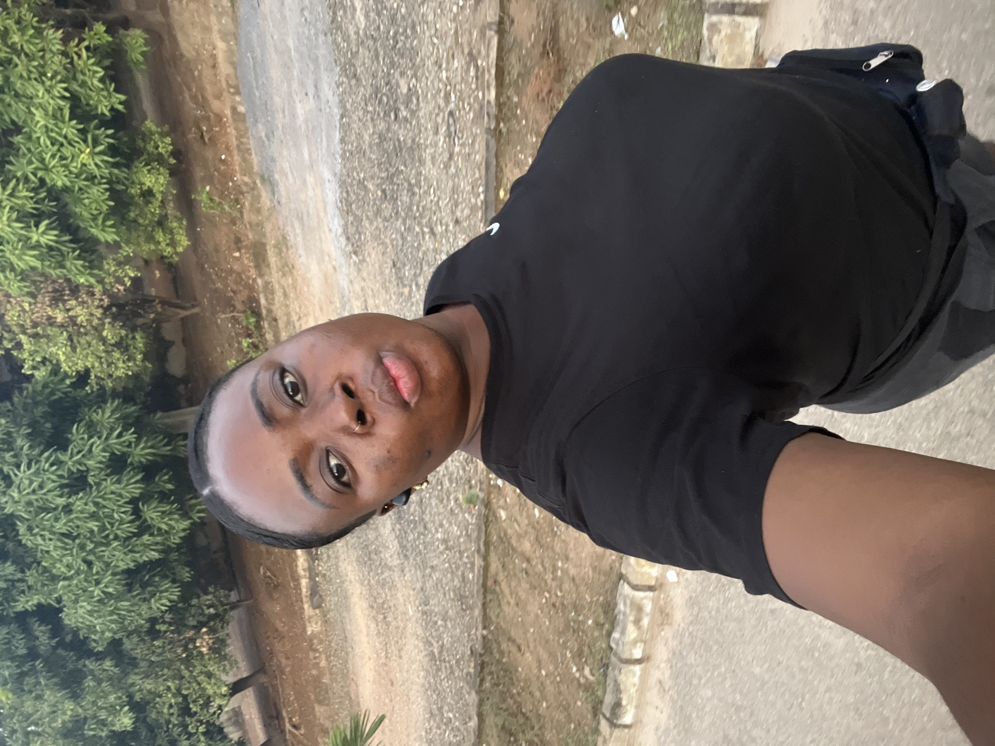 The image is a selfie taken outdoors in what appears to be a residential area in Abuja, Nigeria. A person with brown skin and short black hair is wearing a plain black t-shirt. The background shows a concrete sidewalk, lush green bushes, and a building or wall with a beige color. The location could be a street or a park in a middle-class neighborhood.
