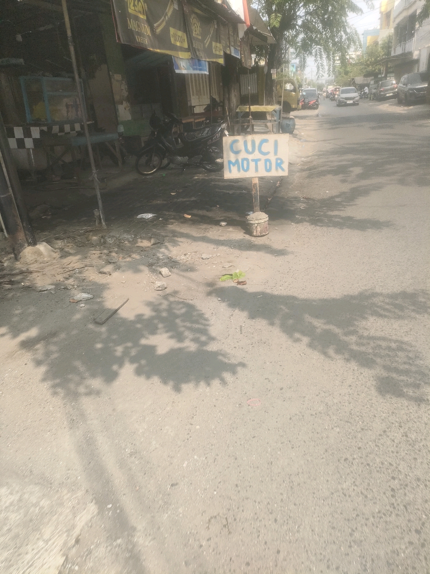 The image depicts a street scene in Surabaya, Indonesia, likely in a commercial or mixed-use area. A makeshift sign with the words "CUCI MOTOR" (motorcycle wash) is prominently displayed in the foreground, indicating a service available. The sign is hand-painted on a white board and mounted on a wooden post. To the left, a dark-colored motorcycle is parked near a weathered storefront or stall. The storefront appears to be constructed with a combination of materials, including what looks like metal sheeting and possibly wood or concrete. Faded banners or awnings hang above the storefront. The street itself is paved and appears to be a public thoroughfare, with vehicular traffic visible in the background, including cars and other motorcycles. The environment suggests a warm, possibly dry climate, with shadows from trees or structures cast across the pavement. The overall impression is of a modest, everyday scene in an Indonesian city.