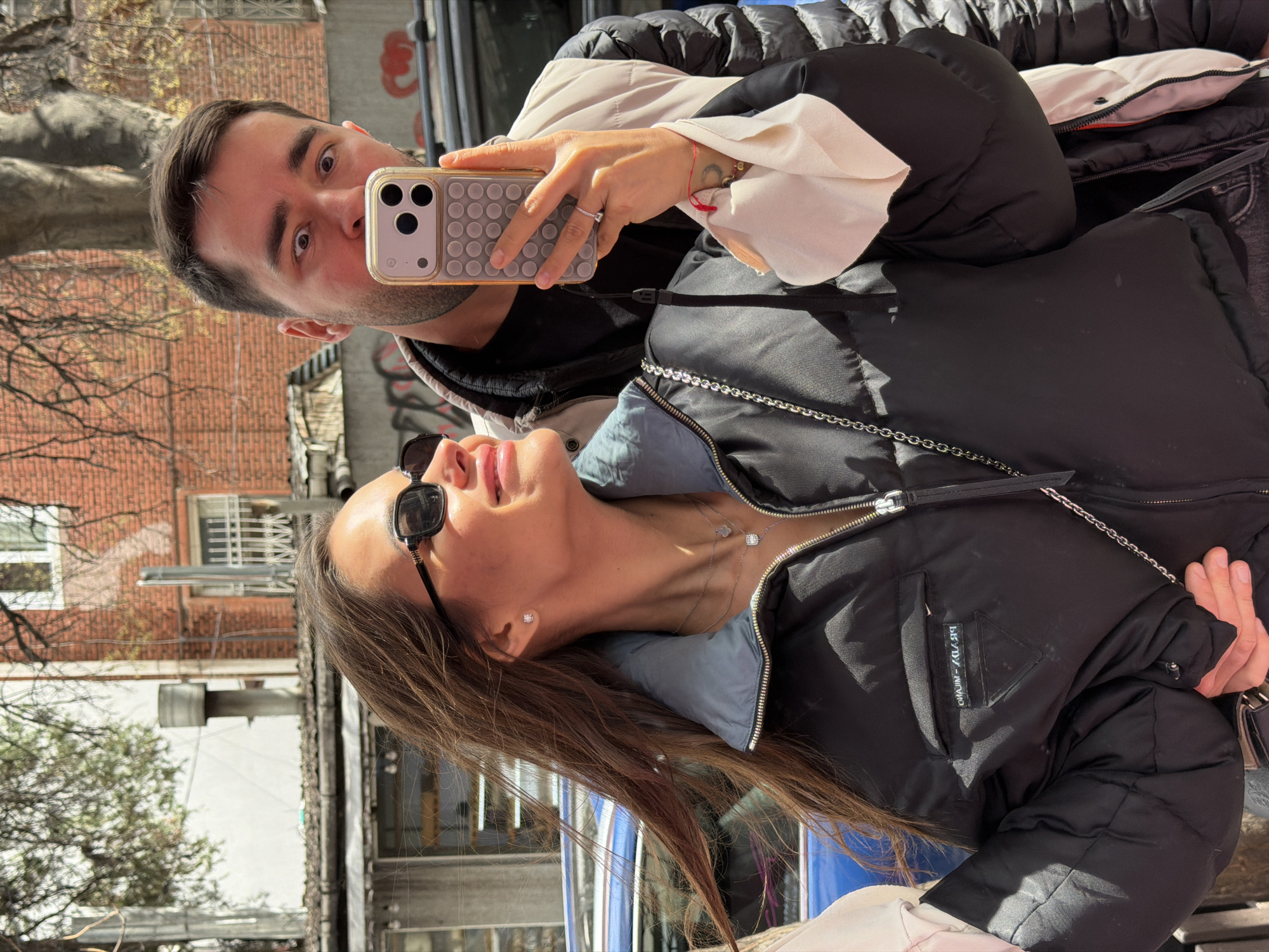 A man and a woman are taking a selfie in the city of Tbilisi, Georgia. The man is holding an iPhone with a speckled case and looking directly at the camera. The woman is wearing sunglasses and a black jacket with a blue collar and a silver necklace. The background includes buildings, trees, and a blue object, possibly a car or bus. The buildings are made of brick and stone and have windows and balconies. The environment appears to be a street with pedestrian traffic.