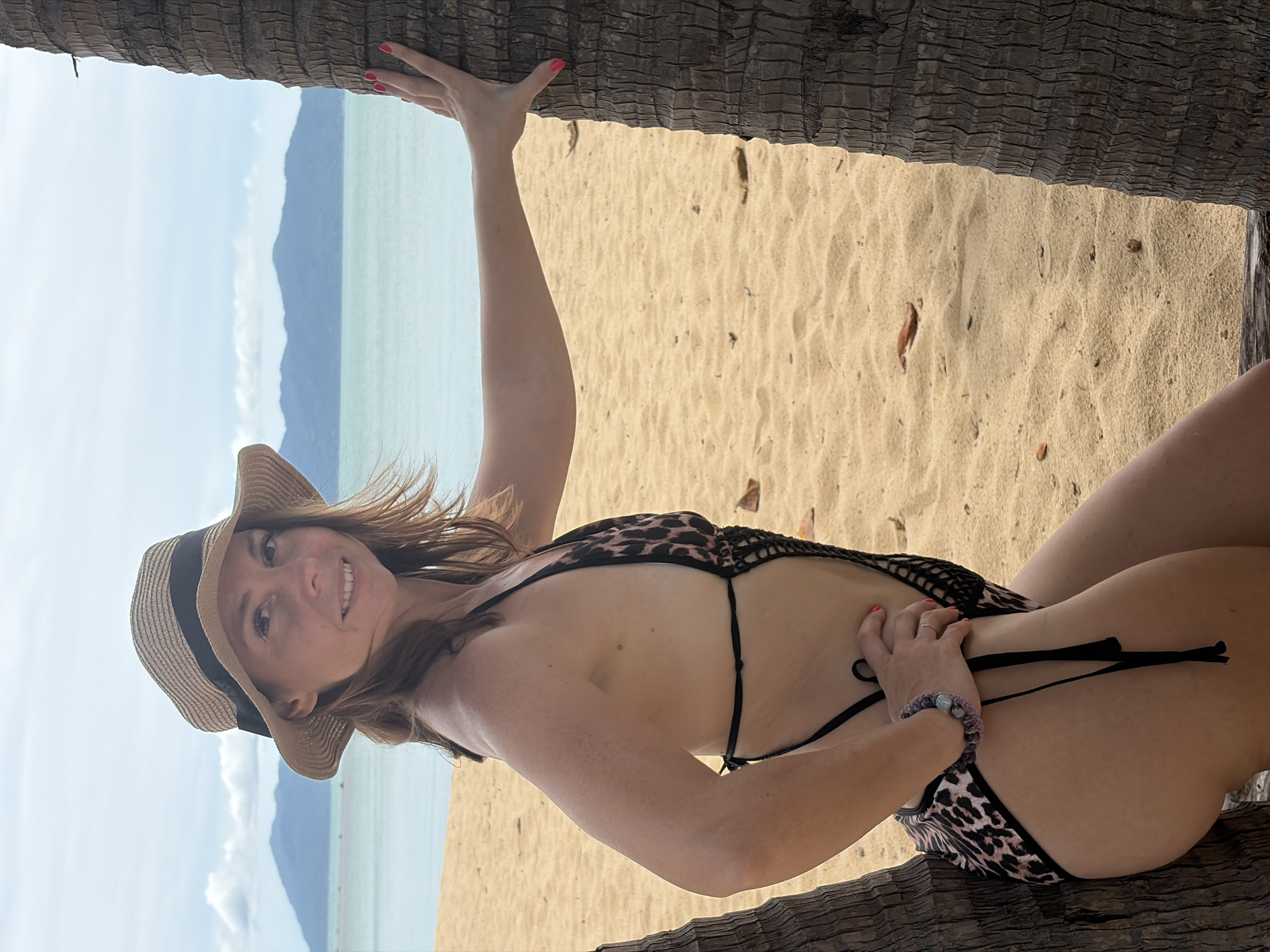 A woman in a leopard print swimsuit and a straw hat poses on a beach in Baan Bo Phut, Thailand. She is leaning against a palm tree, with her left hand resting on its trunk and her right hand on her hip. The ocean and distant mountains are visible in the background. The sand is golden and scattered with fallen leaves. The sky is overcast.