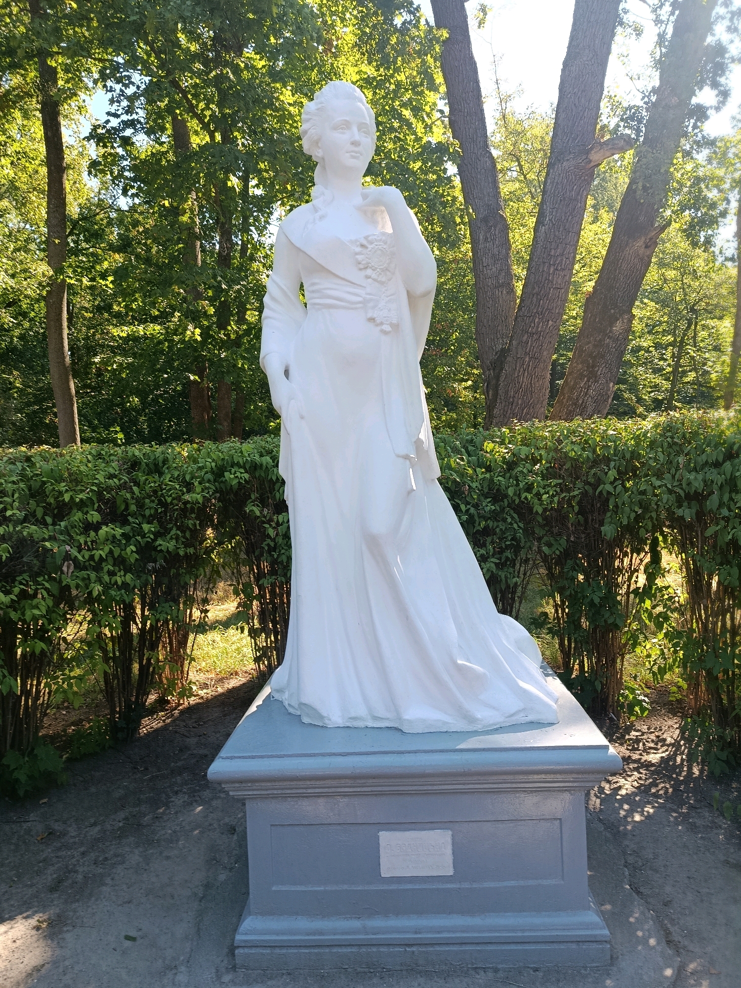 A full-body white marble statue of a woman stands on a grey pedestal in a park setting. The woman is depicted in a formal gown with her hair styled in an updo. She stands with one hand on her hip and the other resting on her chest. Behind the statue, there is a well-maintained green hedge, and behind that, several large trees with dappled sunlight filtering through their leaves. The ground in front of the statue is paved with dark grey material. The base of the pedestal has a plaque with text that is illegible.
