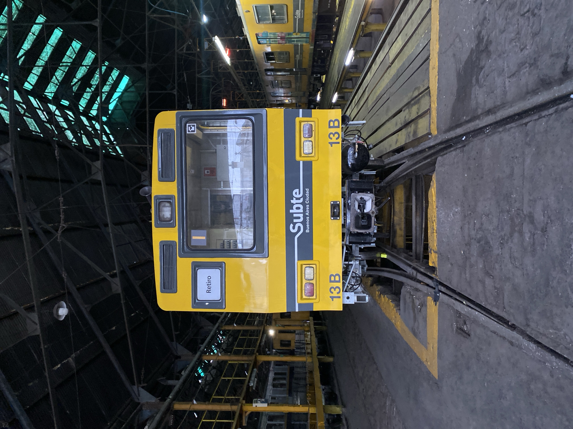 The image captures the front of a yellow and gray Buenos Aires Subte (subway) train, identified as "13 B," positioned on tracks within a maintenance depot in Buenos Aires, Argentina. The train's destination sign displays "Retiro," and the livery includes "Subte Buenos Aires Ciudad." The front of the train features a large windshield, two sets of headlights/taillights, and a central coupling mechanism. The immediate foreground shows a concrete floor with yellow painted lines, leading to a track system with a recessed pit covered partially by wooden planks. The overhead environment consists of a dark, industrial ceiling with exposed steel beams and corrugated metal roofing, featuring multiple translucent panels that allow greenish light to filter in. A single white industrial light fixture hangs from the ceiling. In the mid-ground, other trains are visible: a silver train partially on an elevated platform to the left, and another yellow Subte train parked further down the track to the right. No individuals are present in the scene.