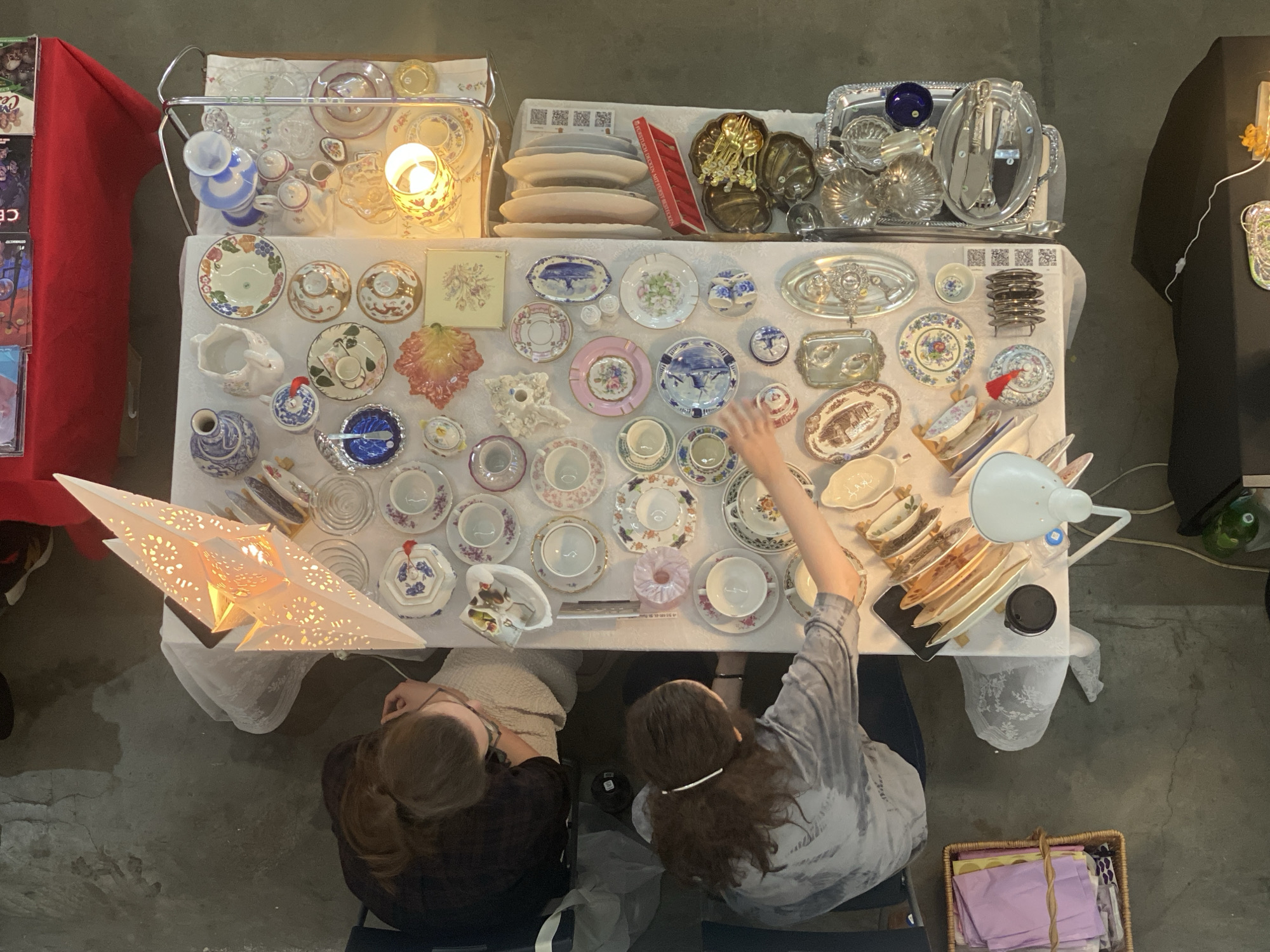 A top-down, indoor shot shows two people sitting at a table covered with a variety of vintage porcelain plates, cups, and other dishware. The table is covered in a white tablecloth. An illuminated star lamp is on the left side of the table. On the right side of the table is a white lamp. One person, wearing a dark shirt, is seated on the left side of the table. The second person, wearing a light-colored shirt, is seated on the right side of the table with their right arm raised. Additional objects, including silverware, are placed on display on elevated surfaces behind the main table. A red drape is on the left of the frame, and a black object is on the right. A basket filled with items is on the lower right of the frame. The location is in the city of Khimki, Russia.