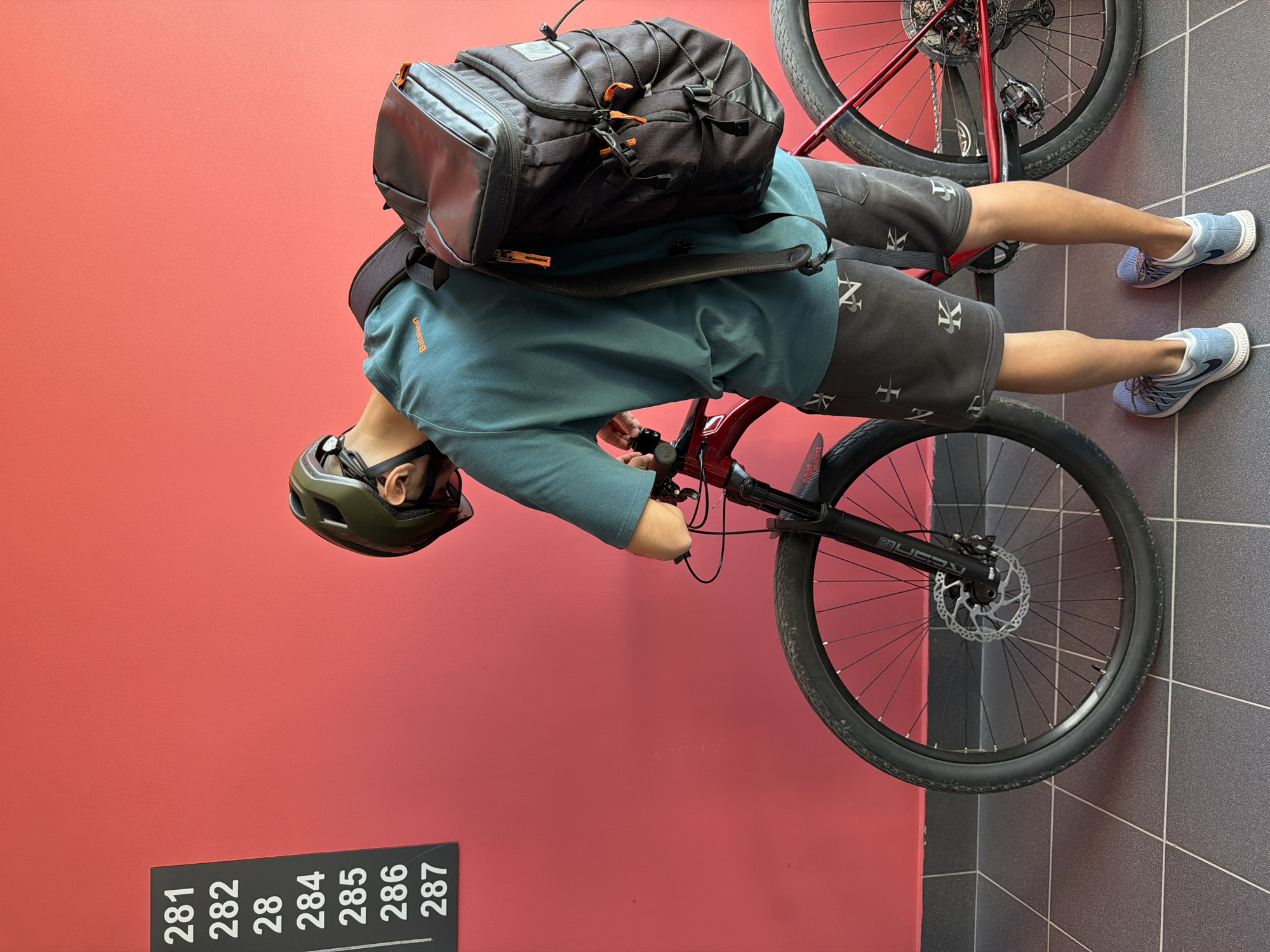 A person wearing a teal shirt, patterned shorts, blue running shoes, and a green helmet is straddling a red bicycle. They have a large black backpack on. The person is near a wall with a red background. On the wall is a sign with the numbers 281, 282, 284, 285, 286, and 287 on it. The floor is made of dark gray tiles with white lines. The location is in Tyumen, Russia.