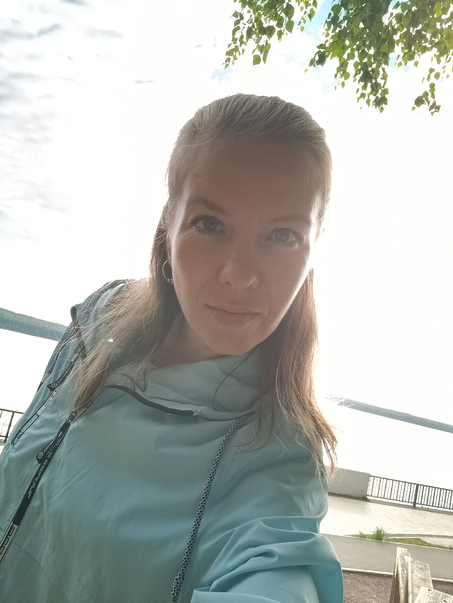 A woman is taking a selfie outdoors, with a body of water and a railing in the background. She has blonde hair pulled back and is wearing a light blue jacket. She has a hoop earring in her left ear. The sky is bright and cloudy, with some green leaves visible in the upper right corner of the frame, suggesting a tree is overhead. The location is the embankment in the city of Chaikovsky, Russia, specifically near the Kama River.