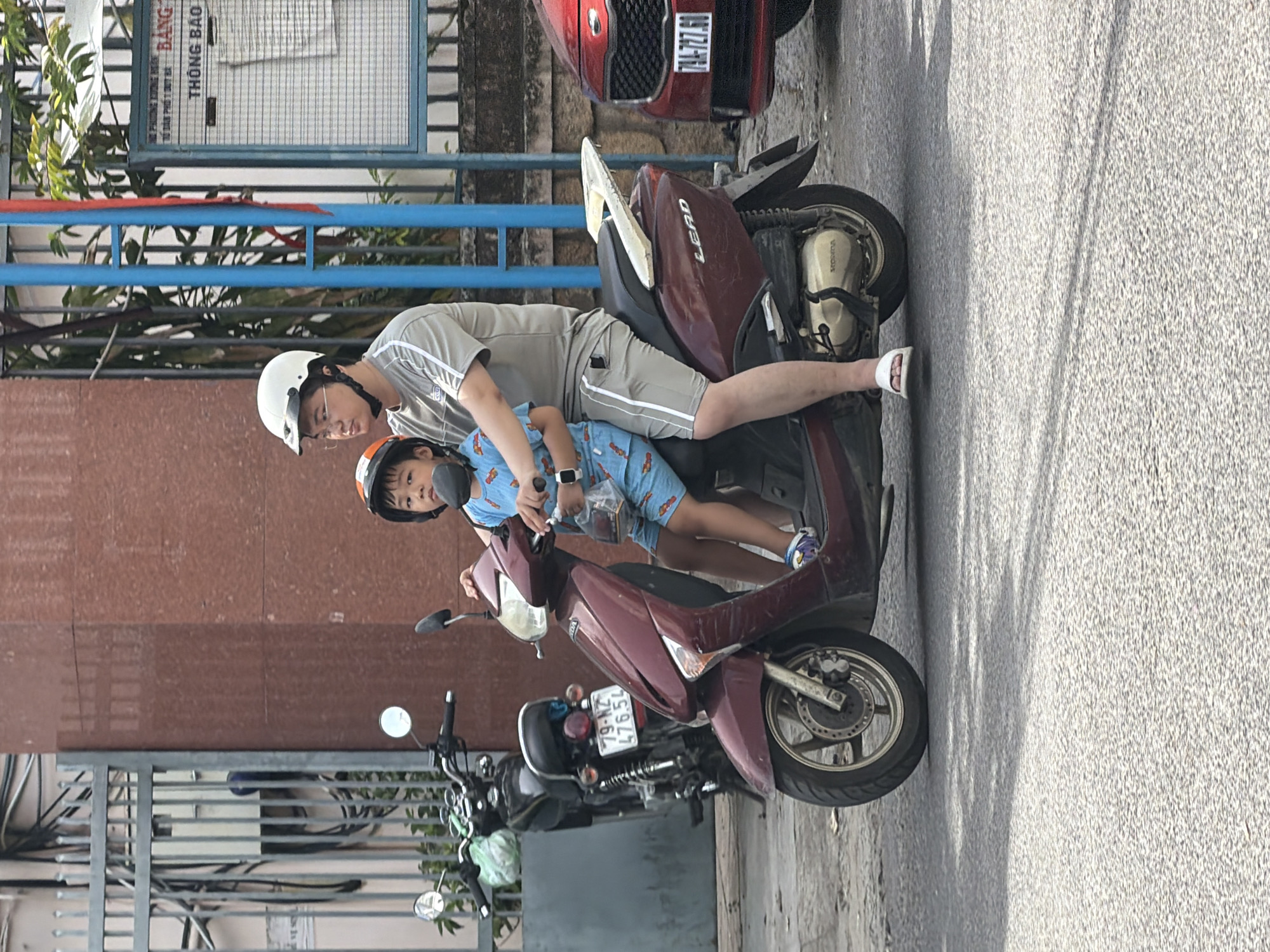A man and a young boy are riding on a maroon Honda Lead scooter on a paved city street in Nha Trang, Vietnam. The man, wearing a light grey t-shirt with white stripes on the sleeves and grey shorts with a white stripe down the side, is driving. He has on a white helmet and glasses. The boy, wearing a blue t-shirt with a cartoon character print and blue shorts with a similar cartoon character print, is seated in front of the man, also wearing a helmet. The boy is holding a plastic bag with a white object inside. The scooter has a license plate that reads "79-NZ 476.54". Behind them, another black motorcycle is parked, with a license plate that reads "79-KZ 476.54". A red car is partially visible on the right side of the image. The background shows a building with a reddish-brown textured wall and a metal fence. There is also a blue metal pole and a bulletin board with Vietnamese text, including the word "THÔNG BÁO", meaning "NOTICE". The ground is asphalt, and shadows are cast across it.