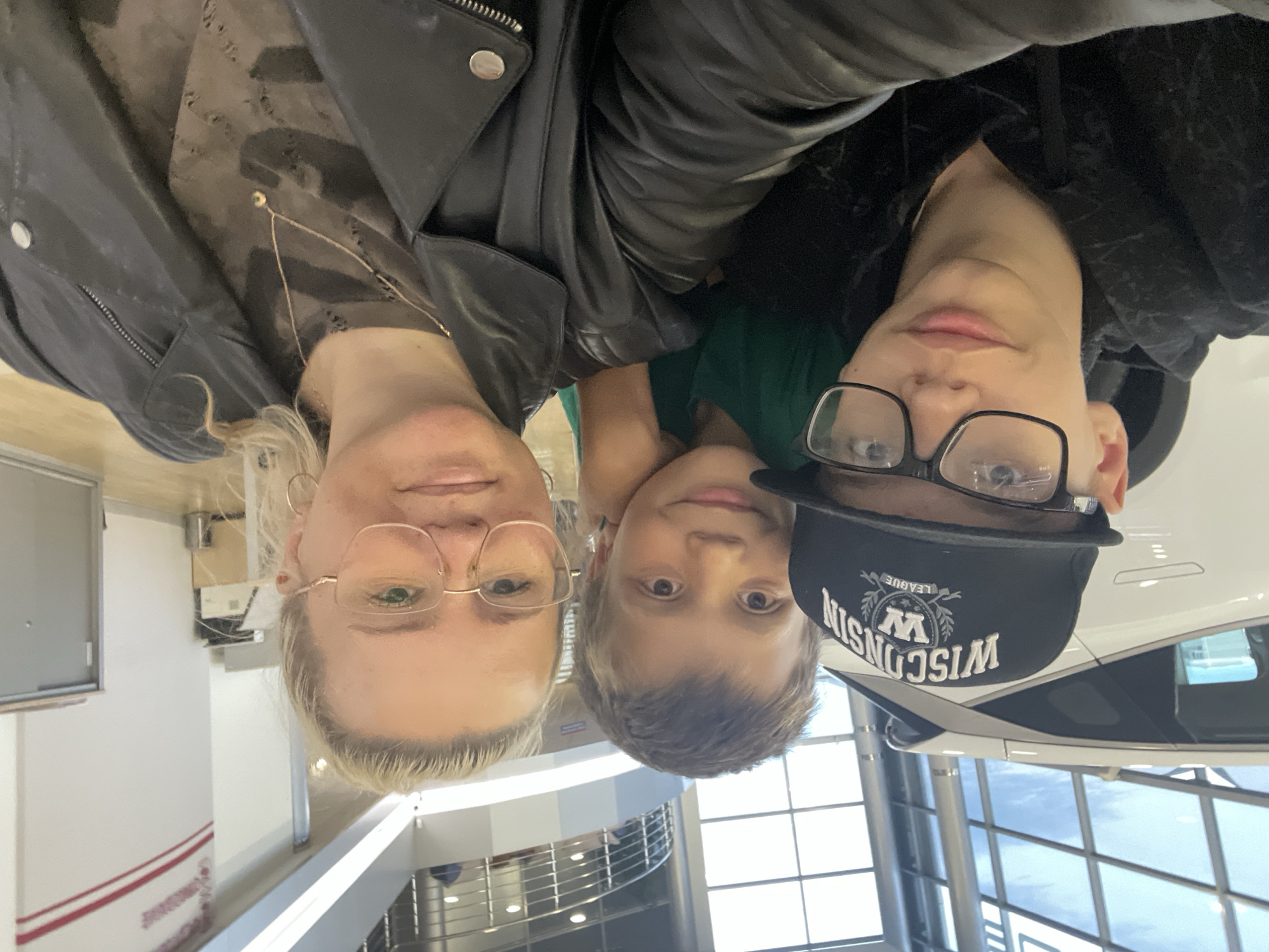 A group of three people, two males and one female, are posing for a selfie in what appears to be a car dealership. The younger male in the foreground, wearing glasses and a black hoodie with a baseball cap, is in the center of the frame. To his right, a boy with short brown hair and wearing a green t-shirt is looking at the camera with a smile. To the right of the boy, a woman with blonde hair and glasses, wearing a black leather jacket, is also looking at the camera. In the background, a white car is partially visible. The environment seems to be indoors, with large windows and a modern architectural design suggesting a commercial space. There are no specific indicators in the image that would allow for a definitive identification of the location within Yekaterinburg, Russia.