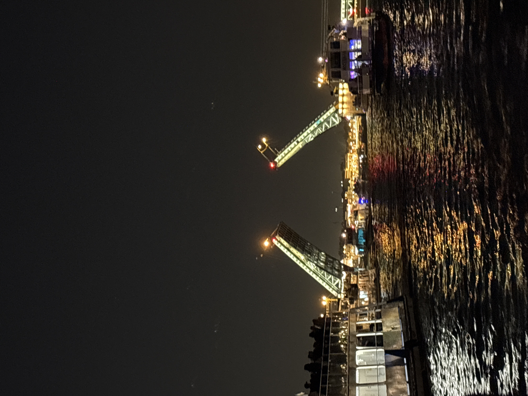 At night, a drawbridge is raised above the Neva River in Saint Petersburg, Russia. The raised sections of the bridge are illuminated with lights that reflect onto the dark water. Buildings and additional light sources illuminate the opposite bank of the river. The sky above is dark, hinting at the late hour. The photograph is taken from a position that suggests being on the river, likely from a boat or embankment.