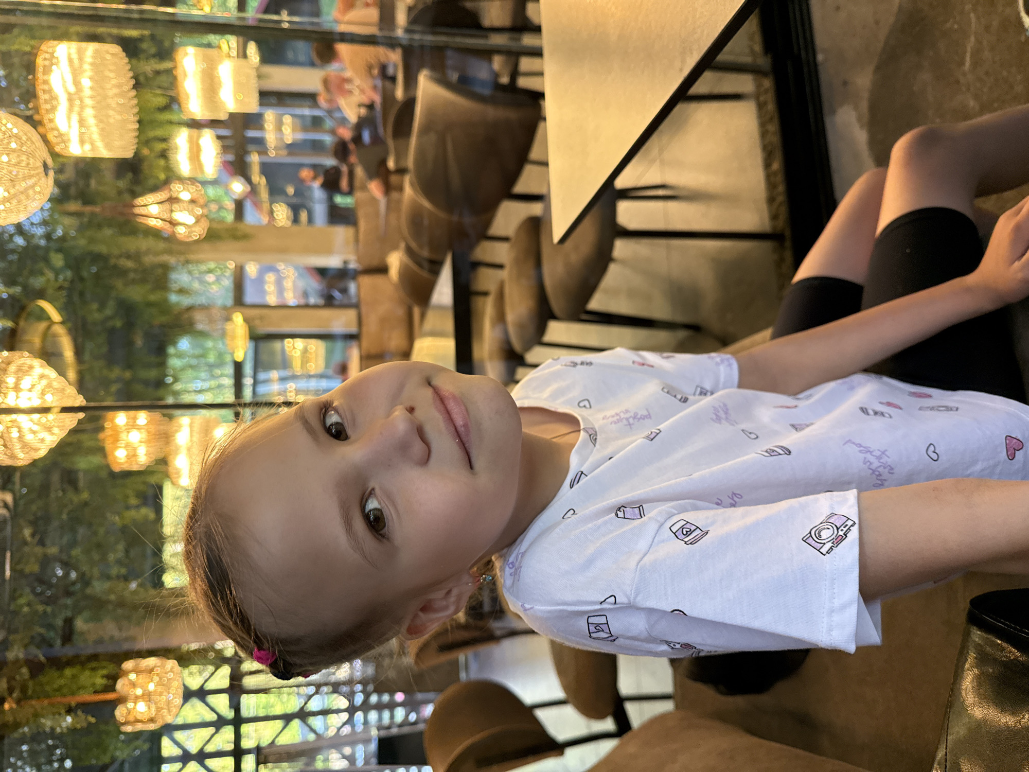 A child is sitting at a table in what appears to be a restaurant or cafe. The child is wearing a white T-shirt with designs, and black shorts or leggings. A table is to her left. Through a glass window are decorative lights and a glimpse of outdoor greenery, suggesting an urban environment. Without explicit location evidence in the provided image, it is impossible to definitively identify the specific location as Izhevsk, Russia.