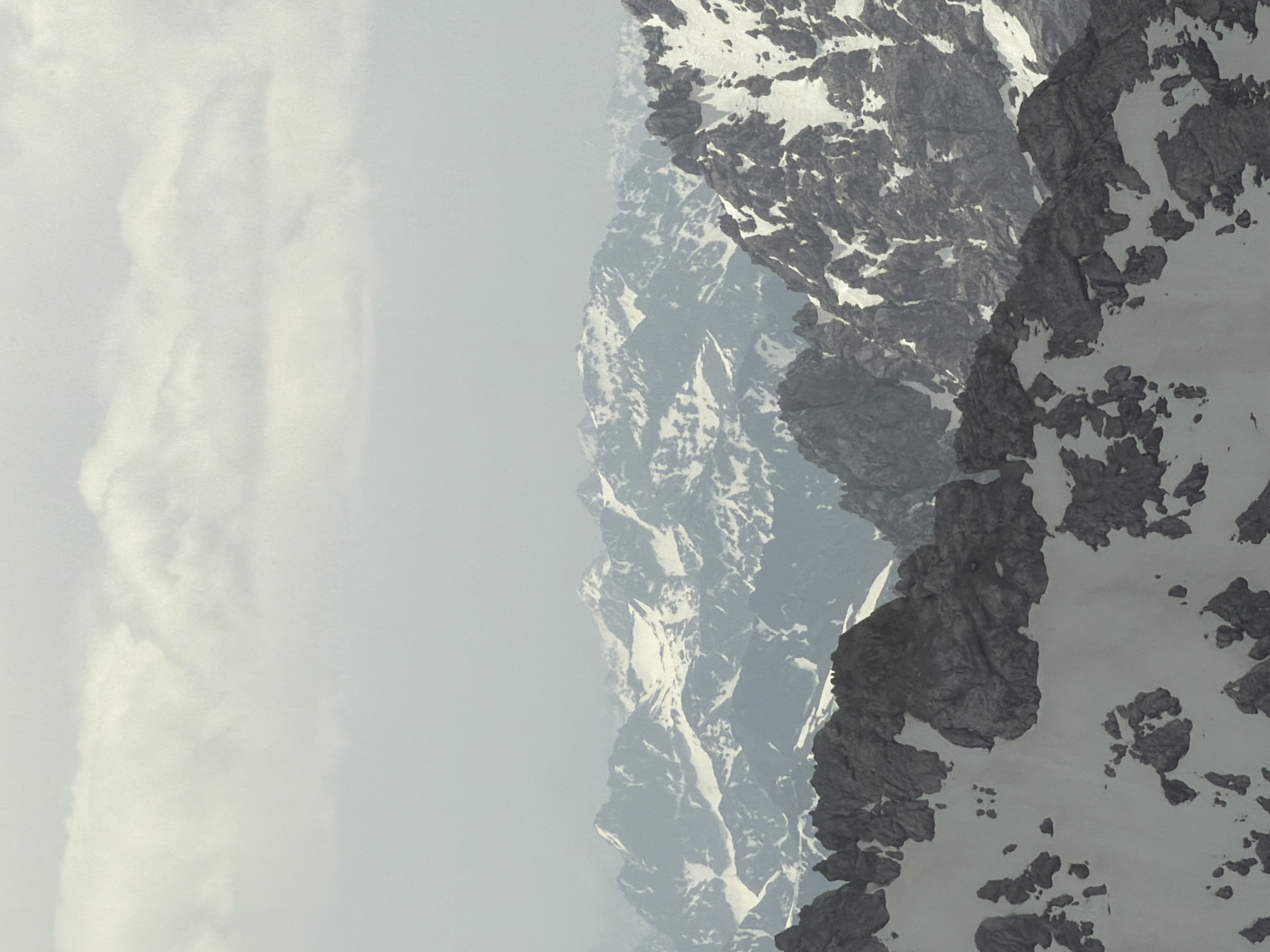 The image displays a portion of a mountain range, partially covered with snow and rock, obscured by mist or fog. The scene appears to be taken on a hazy or overcast day. The mountain range is potentially situated in or near Garmisch-Partenkirchen, Germany, given its alpine environment. The left side of the image is dominated by a light gray, hazy sky. The rocky mountain terrain is heavily interspersed with patches of snow. There are no identifiable people, subjects, or actions evident in the cropped frame.