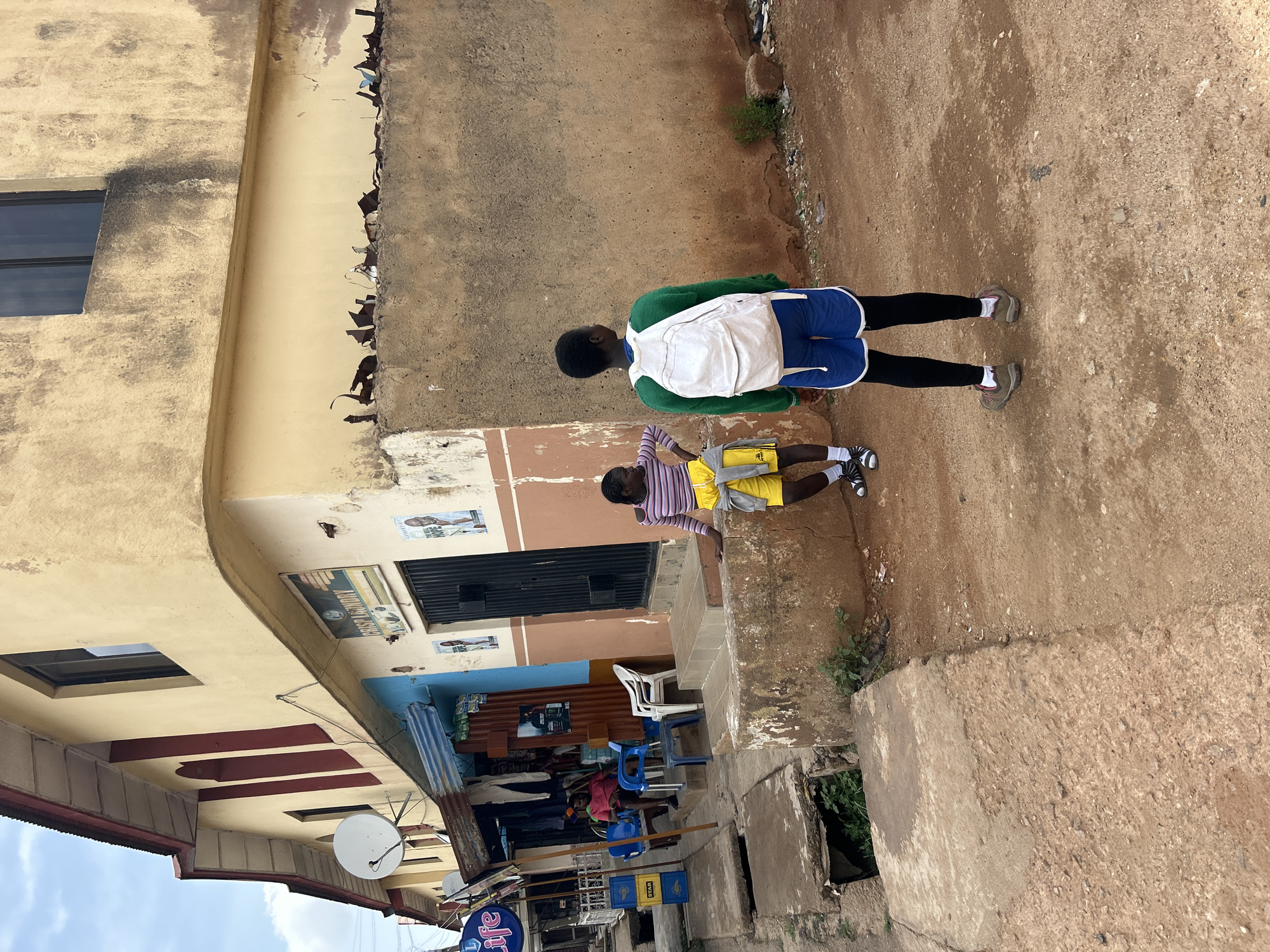 A street in Jos, Nigeria, shows a building with a yellow-beige facade, weathered and chipped. Signs are fixed above the building's entrance. A satellite dish is also mounted. Two young individuals walk along the street: one with a green sweater, blue shorts, and a white backpack; the other in a striped shirt and yellow shorts. Seated under a blue awning are people on blue chairs. Litter is visible on the street.