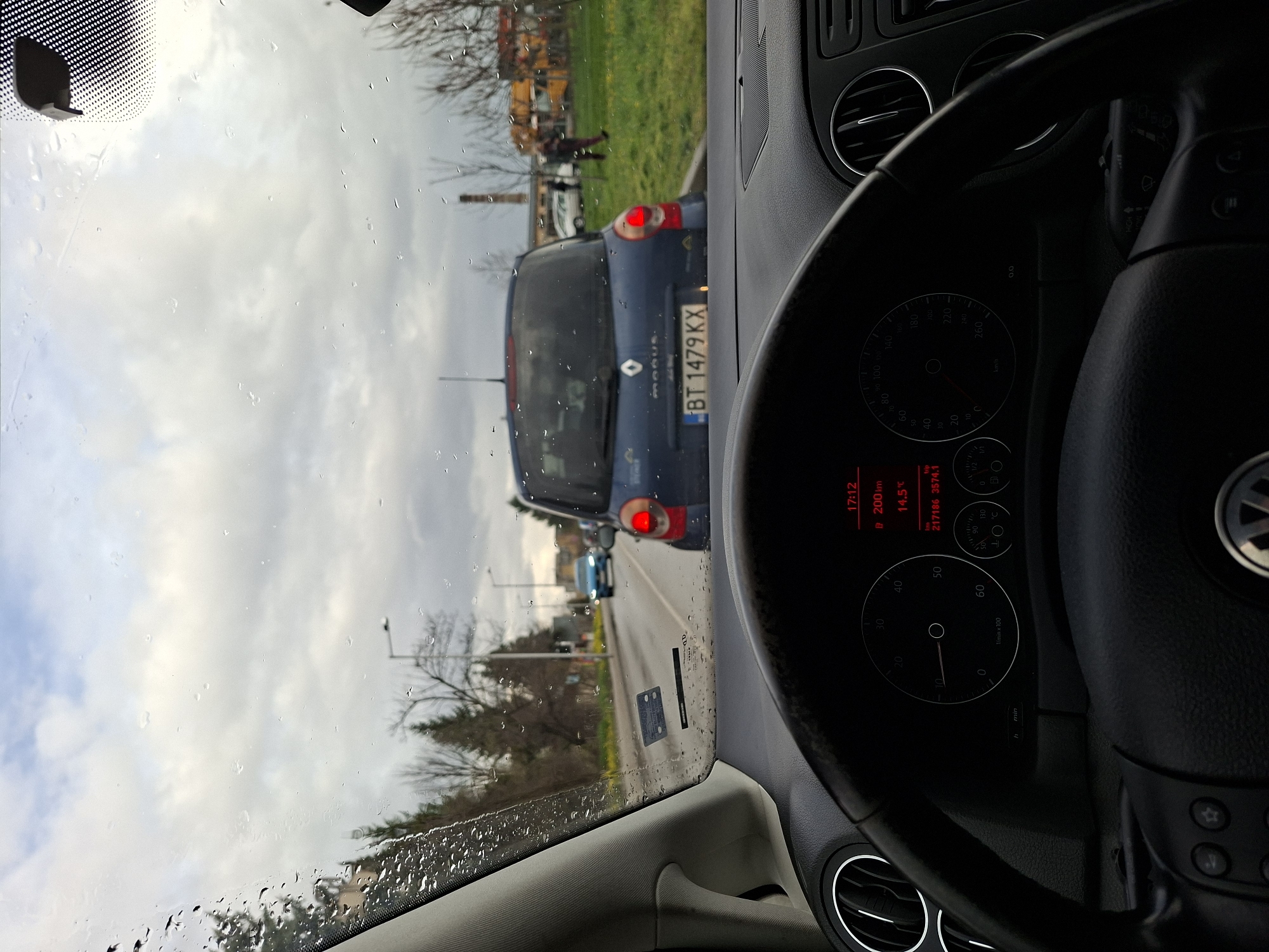 The image shows a view from inside a car, likely a Volkswagen, as seen by the steering wheel and dashboard. The dashboard indicates a time of 17:12, a distance of 200 km, and a temperature of 14.5°C. Through the windshield, it is raining, evidenced by water droplets on the glass. In front of the car, a blue Renault Modus is visible, displaying the license plate "BT 1479 KX," suggesting the location is in Bulgaria, potentially in or around the city of Veliko Tarnovo. Behind the Renault, there's another car, and further in the background, there is greenery and what seems to be a construction site or industrial area. A person is walking on the grass verge. The location seems to be an urban or suburban street with a mix of residential and industrial elements.