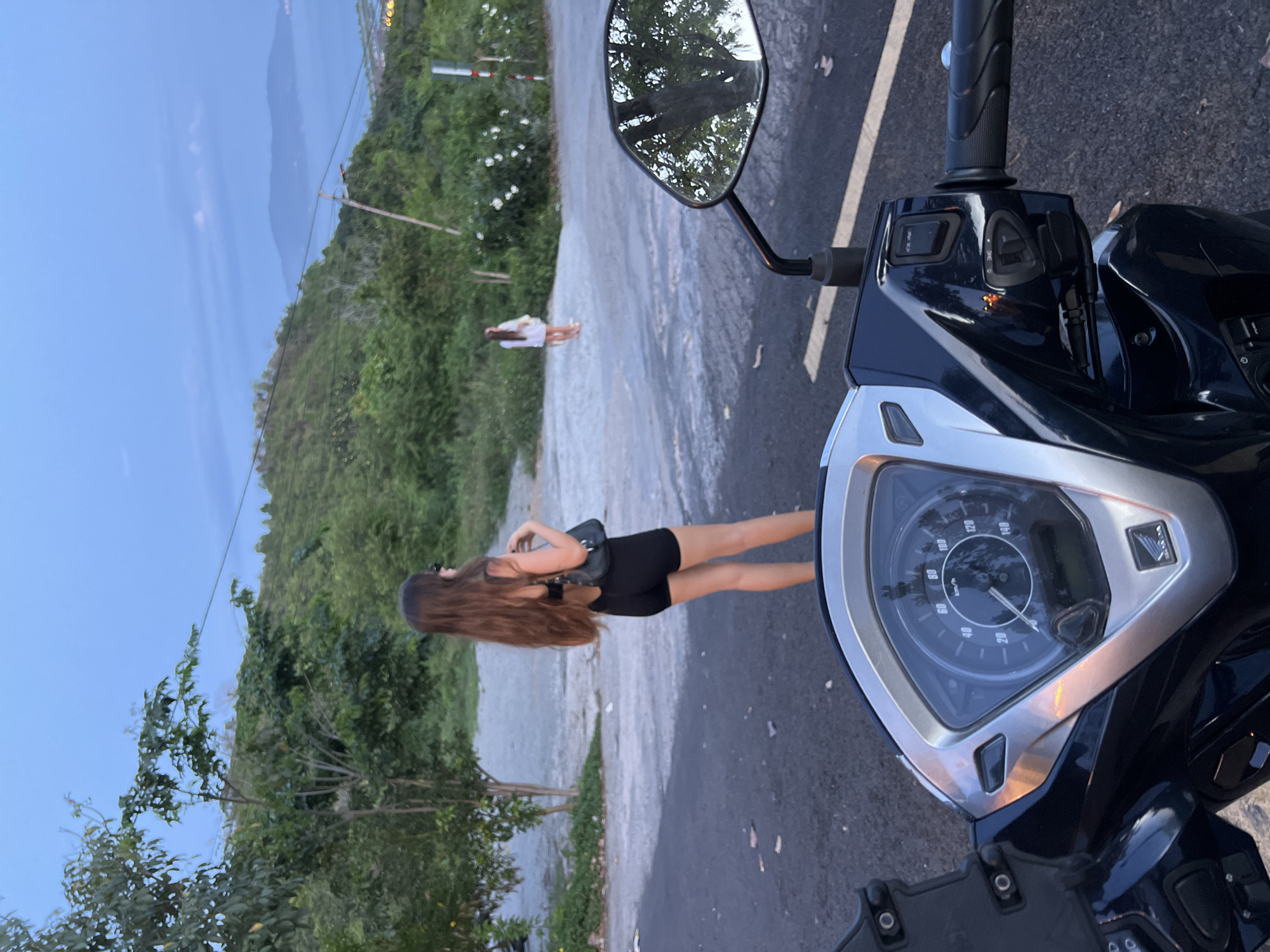 A POV shot from a motorcycle shows a young woman with long brown hair, wearing black shorts and a black top, standing on a paved road with a dirt lot behind her. She is looking away from the camera. In the background, another young woman in a white dress stands near a wall and bushes. The scene appears to be a rural or suburban area with green hills and a body of water in the distance, consistent with the coastal city of Nha Trang, Vietnam. The motorcycle's dashboard is visible in the foreground. The sky is overcast and blue, indicating dusk.