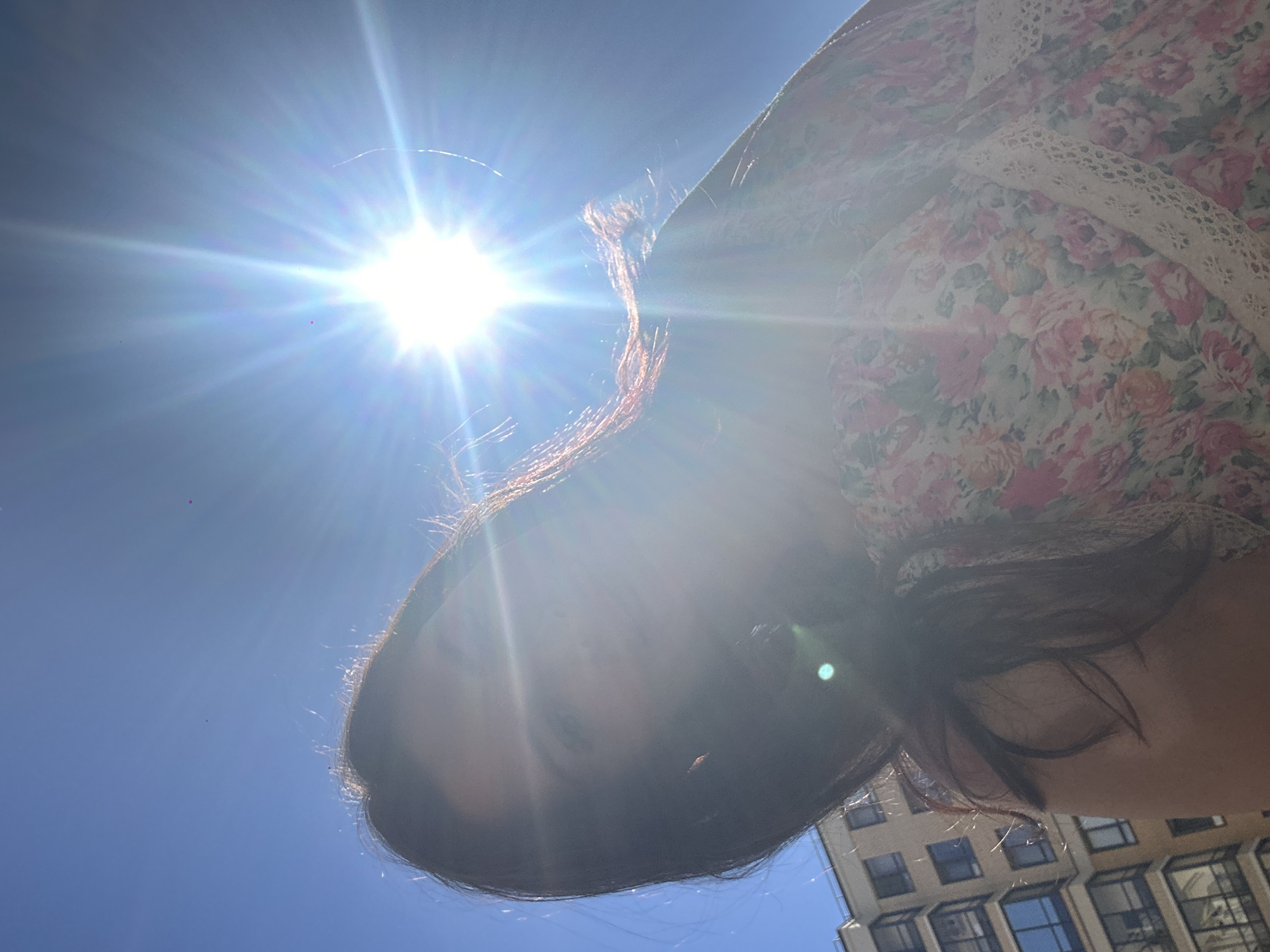 A low-angle, close-up, outdoor shot captures a person's face and upper body against a bright blue sky. The sun is prominently positioned in the upper right quadrant of the frame, emitting strong rays that create a lens flare effect across the image. The person's dark hair partially obscures their face, with strands of hair catching the sunlight. They are wearing a floral-patterned dress with lace trim. In the background, a multi-story building with numerous windows is visible on the left side. The overall impression is that of a bright, sunny day, possibly in an urban environment.