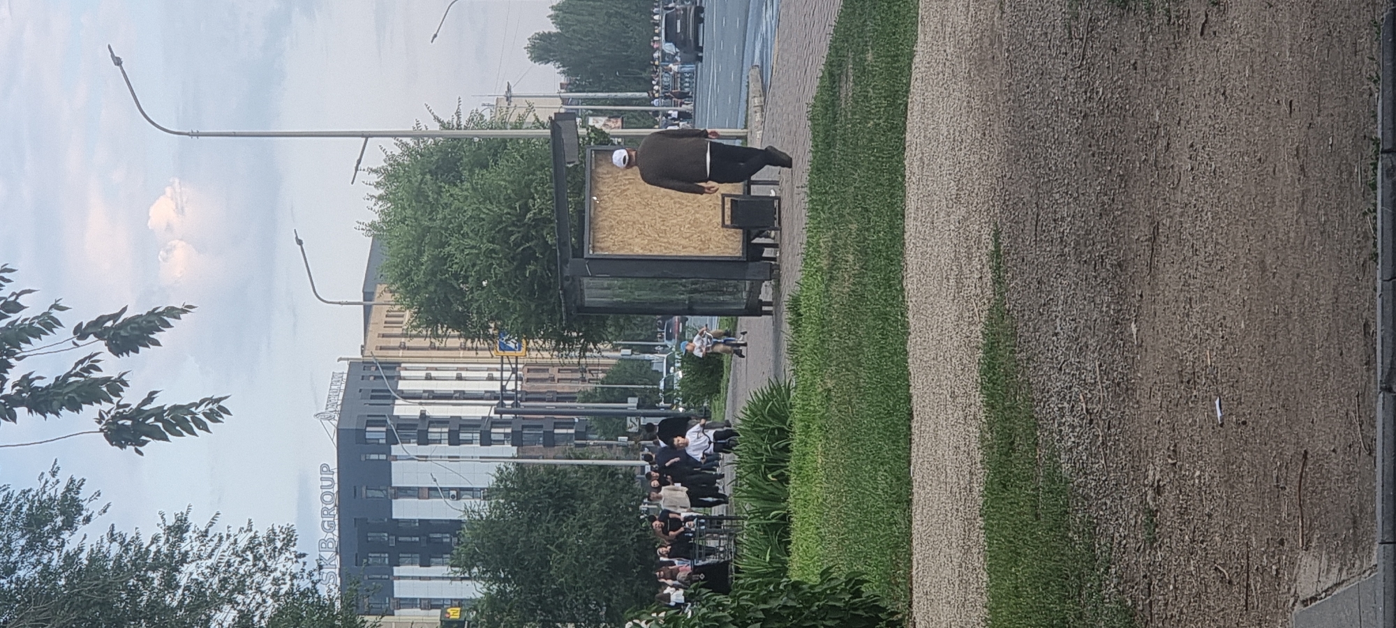 The image depicts a street scene in Aktobe, Kazakhstan, with a modern building in the background labeled "SKB GROUP." In the foreground, a bus stop structure with a wooden panel is visible. Several people are walking on the sidewalk, including a man in a dark sweater and white cap walking away from the camera, and two children riding scooters. A group of people are also seen walking further down the sidewalk. The environment includes trees, a road with vehicles, streetlights, and a cloudy sky. The ground is a mixture of paved sidewalk, gravel, and grass.