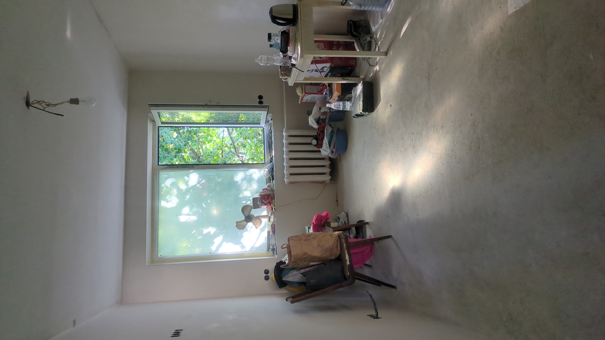 The image depicts an interior room, likely undergoing renovation or being used for storage. The room has plain white walls and a concrete floor. A large window is visible, with one pane open to reveal lush green trees and foliage outside. Sunlight streams through the window, casting distinct patterns on the floor.
In the foreground, a vintage wooden chair is laden with various items, including a large brown paper bag, a dark grey bag, and colorful clothing. To the left of the chair, on the wall, are two black electrical outlets.
In the center of the room, against the wall below the window, is a white radiator. In front of the radiator, on the floor, are several items: a blue plastic basin containing red mugs, a few plastic bags, and other assorted objects.
To the right of the radiator, a white table is set up. On the table are several bottles, a small black electric kettle, what appears to be a pair of glasses, and some boxes. A pair of dark-colored boots rests on the floor near the table. A few other objects, including a toolbox and a large plastic water bottle, are scattered on the floor in front of and to the right of the table.
A small, vintage electric fan is positioned on the windowsill, near the left side of the window.
The room appears to be located in Chișinău, Moldova, based on the provided context. The overall impression is one of disarray, with items accumulated in a functional, though not aesthetically organized, manner.