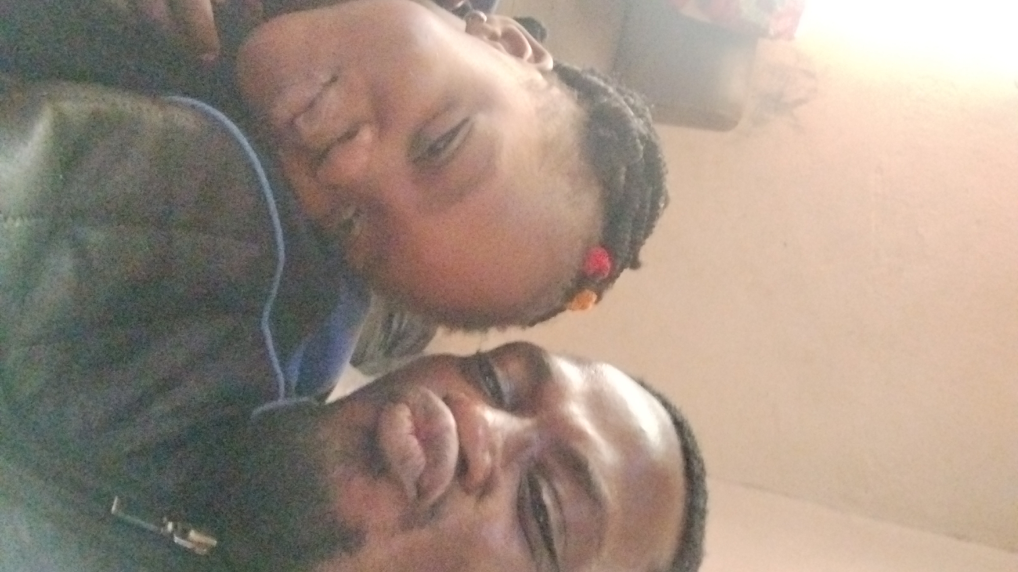 The image shows a close-up, overhead shot of two people, likely a parent and child, with their faces turned towards the camera. The lighting is bright, suggesting it might be daytime, possibly indoors near a window or with artificial lighting. The background appears to be a light-colored wall, perhaps in a home setting.
The person in the foreground, closer to the camera, is an adult with dark skin. Their eyes are open and looking towards the viewer, and their mouth is slightly parted. They are wearing dark clothing, and a metallic object, possibly a clasp or buckle, is visible on their shirt.
The second person, positioned above and slightly behind the adult, is a child, also with dark skin. The child's eyes are closed or nearly closed, suggesting they are sleepy or resting. Their hair is styled in braids with colorful beads, red and orange, at the ends. A light blue string or cord is visible on the adult's clothing, close to the child's head. The overall mood seems to be intimate and caring, possibly a moment of rest or affection between the two individuals. No text is visible in the image.