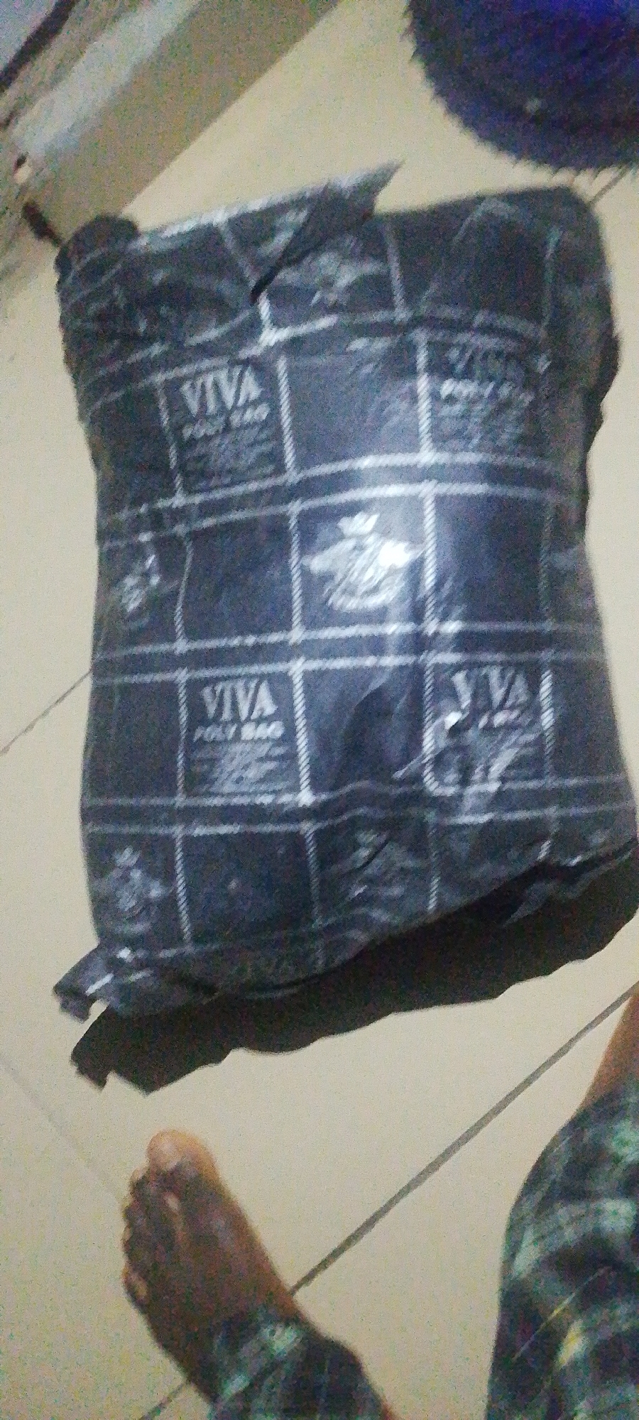 In Jauro Yinu, Nigeria, a parcel wrapped in black plastic with a repeating grid pattern and the "VIVA POLY BAG" logo is positioned on a tiled floor. The parcel appears to be somewhat compressed and irregularly shaped. A person's foot, wearing dark patterned clothing, is visible in the lower left corner of the frame, near the parcel. The background is indistinct, suggesting an indoor or covered outdoor environment with neutral-colored surfaces.