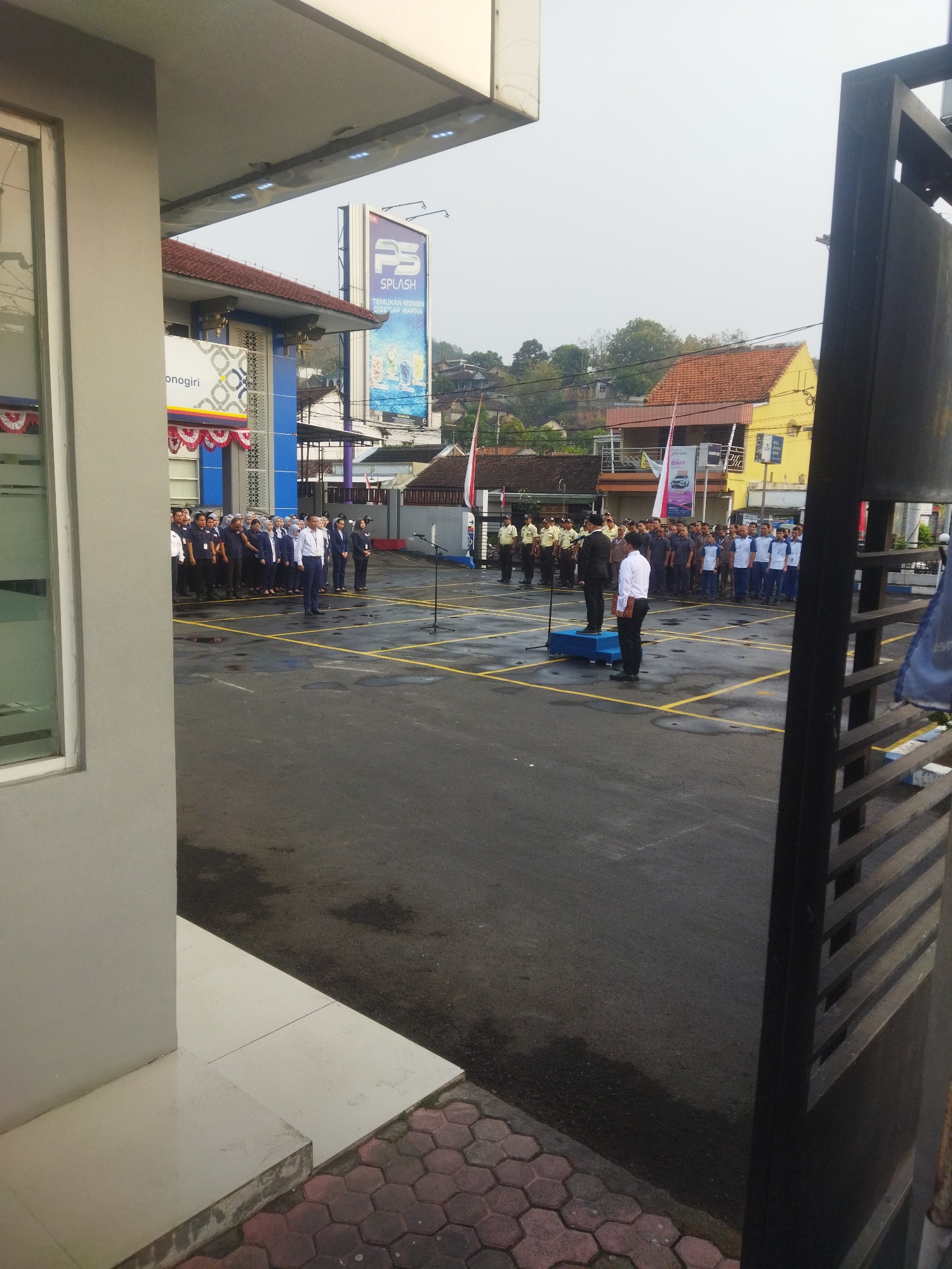 The image depicts an outdoor gathering of people in front of a blue and white building in Giripurwo, Indonesia. A group of men, some in uniform and others in formal attire, are standing in formation on a paved area marked with yellow lines. Two men are positioned on a raised platform at the front of the gathering, with one speaking into a microphone. The building in the background features a red-tiled roof and a prominent billboard advertising "SPLASH." Trees and residential structures are visible on a hillside behind the building. The ground appears wet in some areas, suggesting recent rain. A black metal fence or gate is present in the foreground on the right side of the image. The overall scene suggests a formal event or ceremony taking place outdoors.