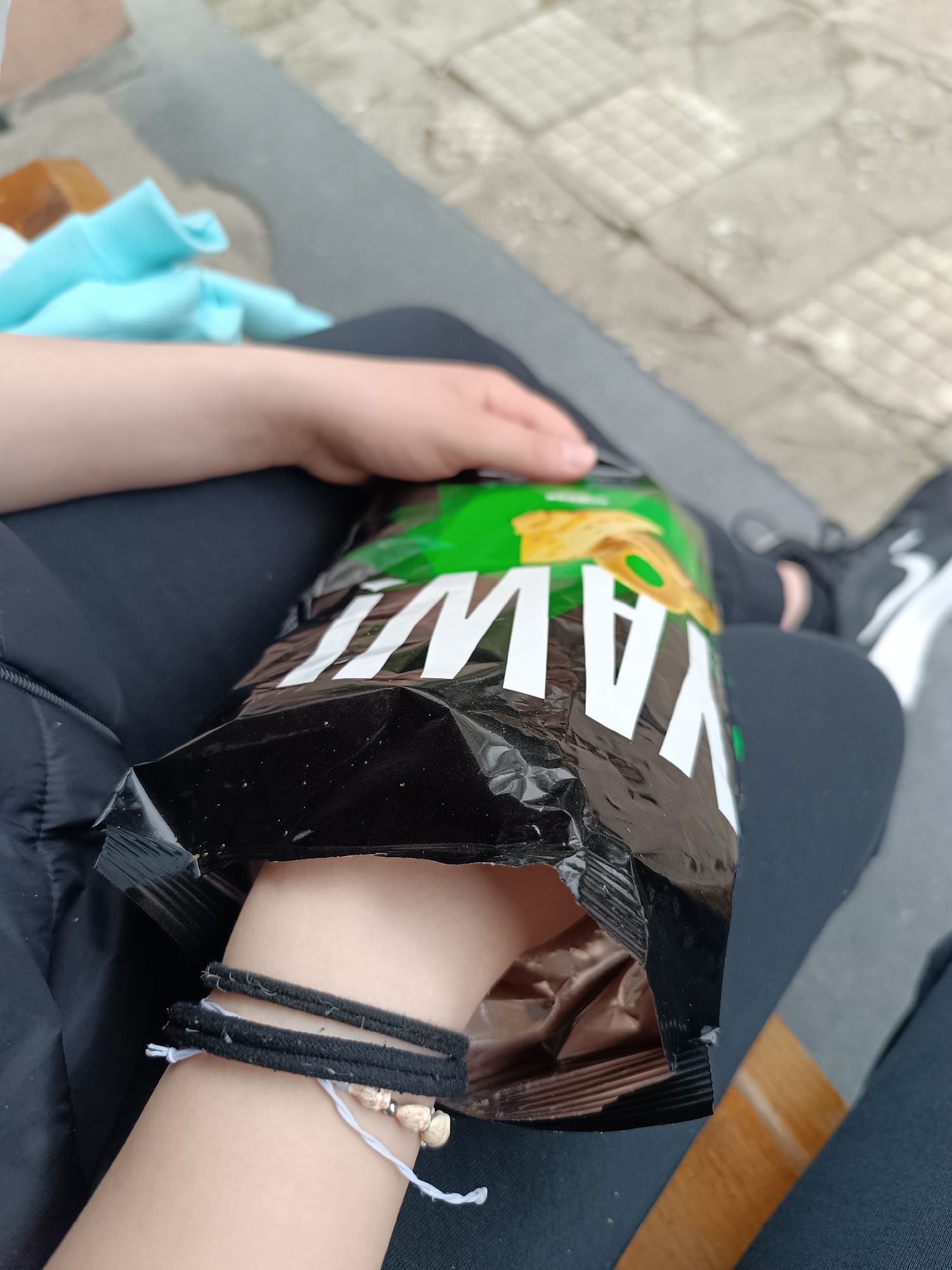 The photograph presents a close-up, high-angle view of a person sitting outdoors and snacking. The subject's arm, hand, and legs are visible, dressed in dark clothing, including what appears to be black trousers and a jacket. A light blue garment is partially seen in the upper left background. The person's right hand, adorned with multiple bracelets (two black fabric bands and a white string bracelet with light-colored beads or shells), is reaching into a black and green bag of chips.
Visible on the bag in large white letters is the reversed text "YAW", which is likely the end portion of the "LAY'S" brand logo seen upside down. The immediate setting features a textured ground surface, composed of light-colored, possibly tiled or paved, sections, alongside a wooden surface in the lower right, suggesting an outdoor public space. The lighting indicates it is daytime. The image captures a casual moment of eating in Pleven, Bulgaria.