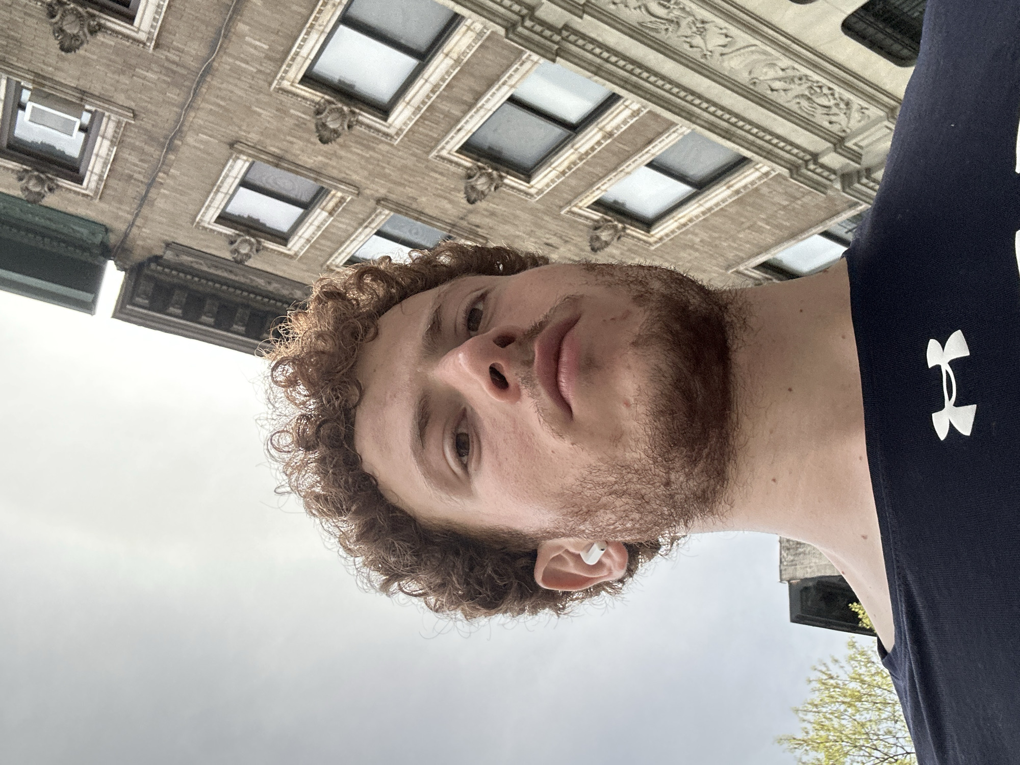 A young man with curly brown hair and a light beard is captured in a low-angle shot, looking slightly upwards. He wears a dark blue t-shirt with a white Under Armour logo prominently displayed on his right shoulder/chest area. A white earbud is visible in his right ear. His expression is neutral, and his gaze is directed slightly off-camera.
The scene is outdoors, set in an urban environment consistent with New York, United States of America. Behind and above the man, a multi-story building with a brick facade dominates the background. The building features numerous windows with ornate, light-colored decorative frames and sills, suggesting classical architectural influences. Intricate carvings and cornices are visible along the building's upper sections and above the windows. The sky overhead is overcast, indicating a cloudy daytime setting. A hint of a tree with nascent green leaves is visible in the bottom right corner of the frame. The overall perspective is from directly below the subject, looking up towards the building and sky.