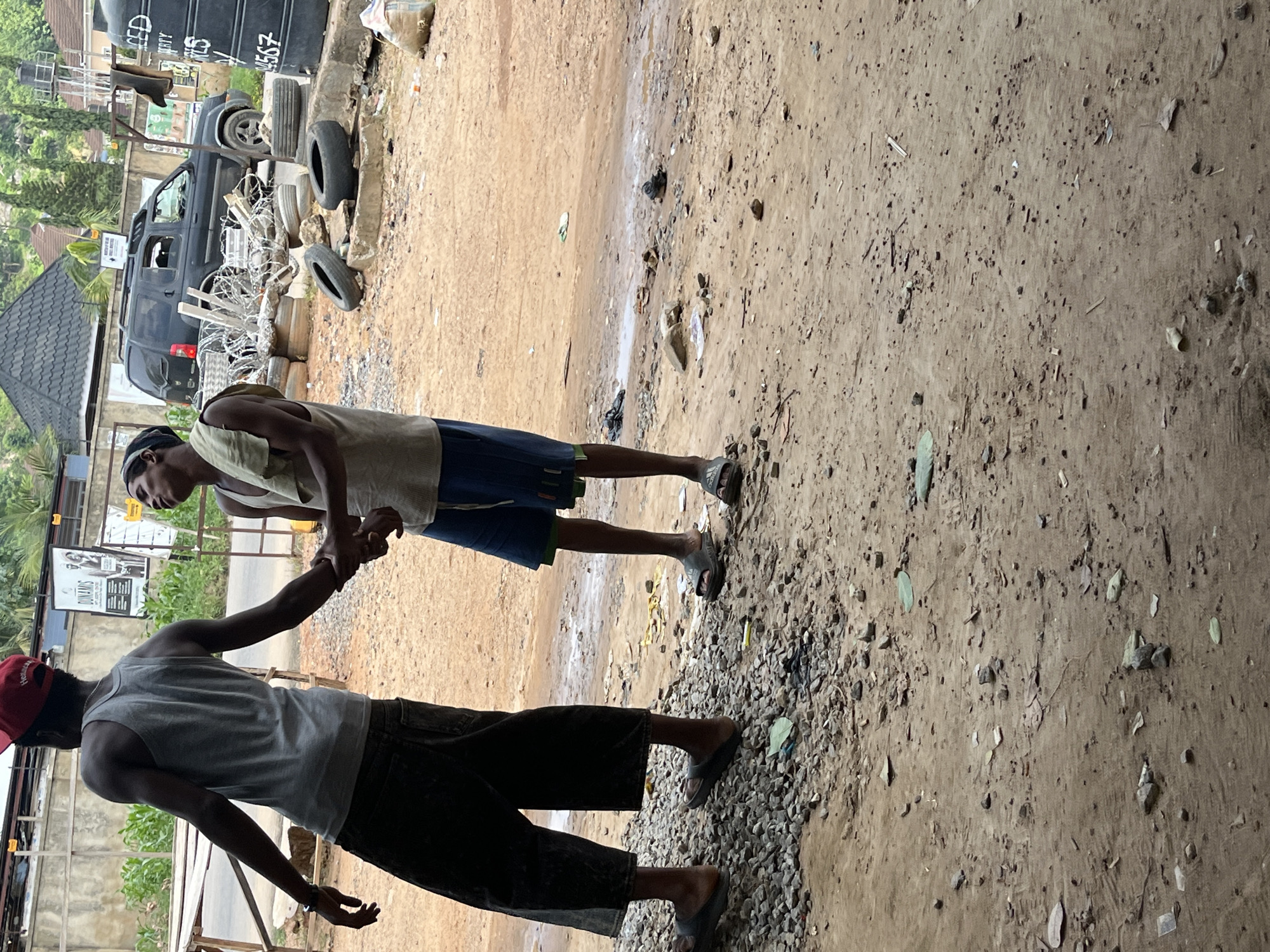 The image shows two men standing in what appears to be an outdoor, possibly informal marketplace or workshop area in Abuja, Nigeria. The ground is a mix of dirt, gravel, and some scattered debris. In the background, a dark-colored SUV is parked near a large black water tank. Tires and what looks like barbed wire are also visible, suggesting some sort of security or storage. A building with a dark, sloped roof and windows is partially visible on the left, along with some greenery.
The two men are interacting, with one man offering his hand to the other, who is shaking it. The man in the foreground is wearing a grey tank top and dark shorts, with a red baseball cap. The man behind him is wearing a light-colored tank top and blue shorts. They are both wearing sandals. The overall impression is of a casual, possibly business-related encounter. The lighting suggests it is daytime, with no strong shadows indicating early morning or late evening. The weather appears to be clear. There is some text visible on the water tank in the background, including "LED" and a sequence of numbers that looks like "94567". There is also a sign with text, possibly advertisements, in the background.
