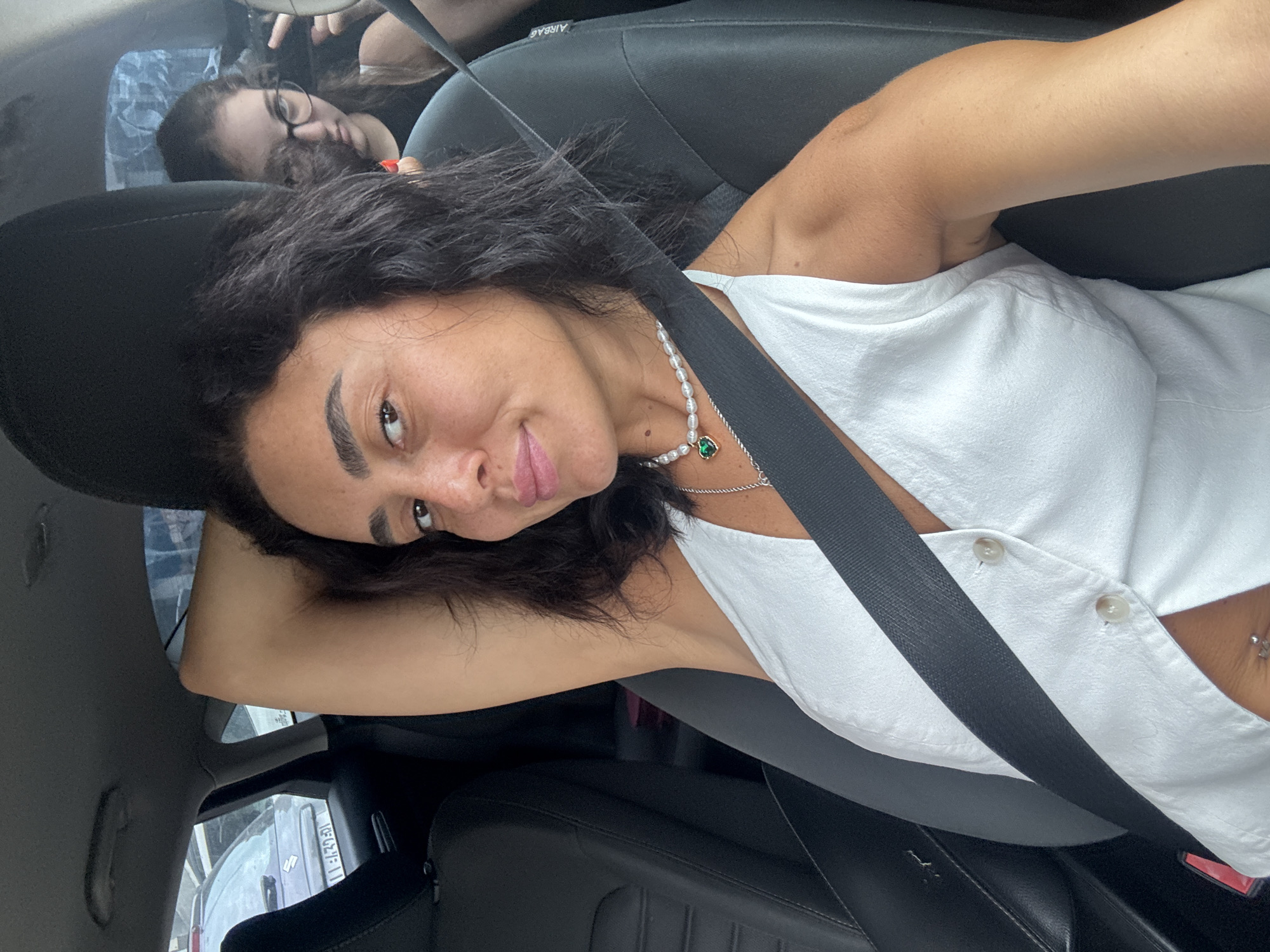 The image shows two females in the front seats of a car, likely in Tbilisi, Georgia. The primary subject, in the driver's seat, is a woman with dark, wavy hair, wearing a white top and a pearl necklace with a green heart pendant. She is looking towards the camera with her left arm raised behind her head. In the passenger seat, partially visible, is a younger female with dark hair and glasses, looking towards the driver. The interior of the car is visible, including a black seatbelt across the driver's chest and the black leather of the car seats. Outside the car windows, other vehicles and a building are discernible, suggesting an urban environment.