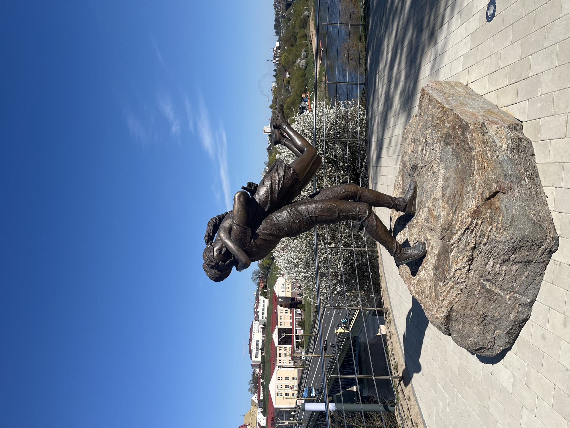 A bronze sculpture depicting two embracing figures, presumably a man and a woman, stands on a large, rough natural rock pedestal on a sunny day in Hrodna, Belarus. The figures, rendered in a realistic style, show one individual seemingly lifting or supporting the other, who is leaning back in a tender embrace. They are dressed in casual attire, including shoes and trousers.
The sculpture is positioned on a grey paved walkway. Behind it, a metal railing separates the viewing area from a landscape featuring a river and the city beyond. The river flows through a valley, and its banks are dotted with various buildings, some with red roofs, and numerous trees, several of which are in bloom with white flowers, suggesting springtime. In the distance, across the river, more buildings and what appears to be an industrial chimney emitting smoke can be seen.
The sky is a clear, vibrant blue with a few faint cirrus clouds. Strong sunlight from a high angle casts prominent shadows of the sculpture and the railing onto the pavement, indicating it is likely midday or early afternoon. The scene evokes a sense of romance and peacefulness amidst an urban riverside setting. There is no visible text in the image.