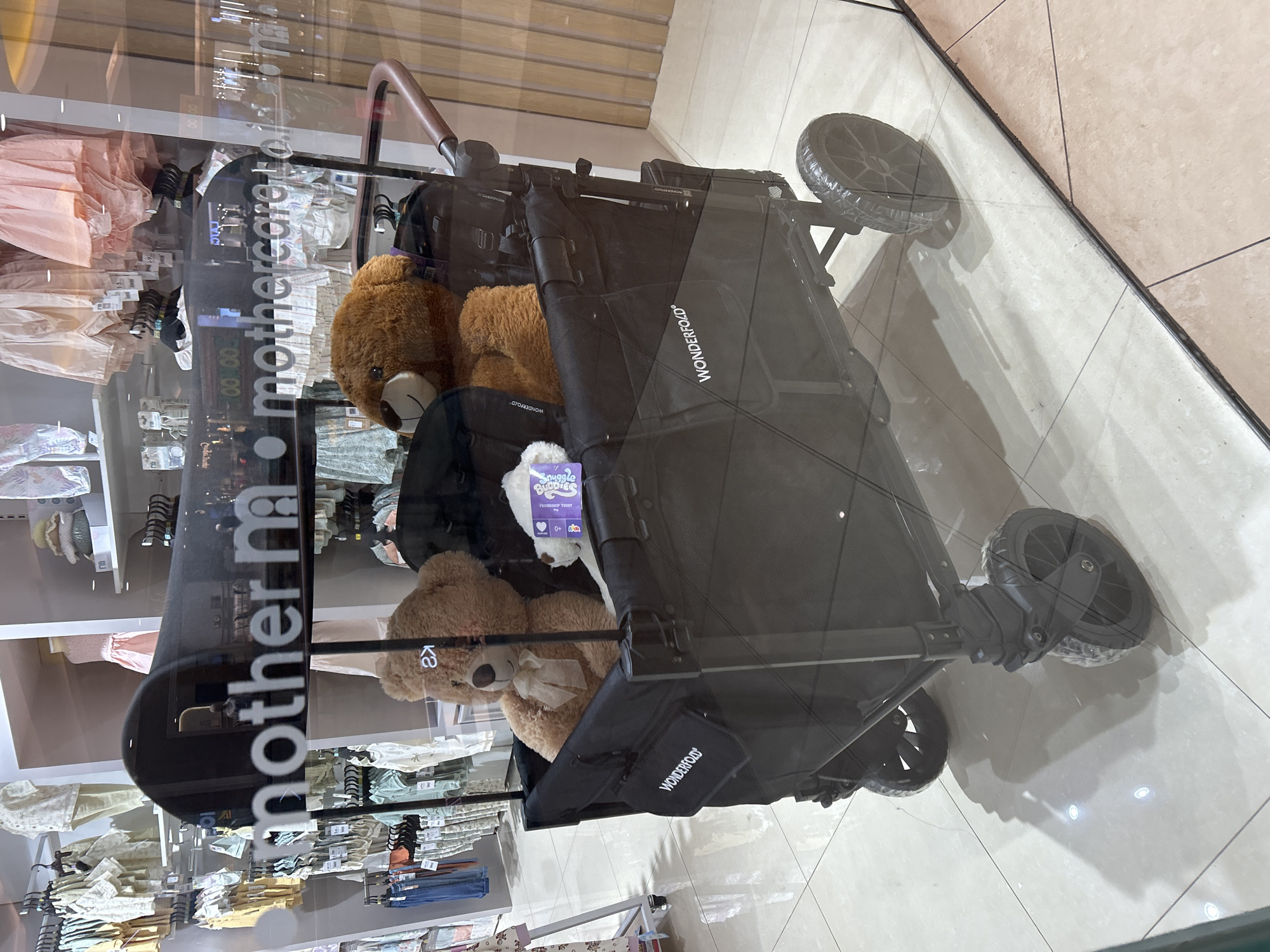 This is a photograph of a black Wonderfold wagon inside a store, viewed through a glass window. The wagon contains two teddy bears, one large brown bear with a bow and a smaller white bear. The word "WONDERFOLD" is visible on the side of the wagon. In the reflection on the glass, the words "mothercare" are prominent. Behind the wagon, the store appears to be a children's clothing store with racks of baby clothes on display. The floor is tiled with a light-colored pattern. The photograph is taken from a low angle, looking upwards into the wagon and the store. The lighting suggests an indoor retail environment. The location is identified as Makassar, Indonesia, implying this is a scene from a Mothercare store in that city.
