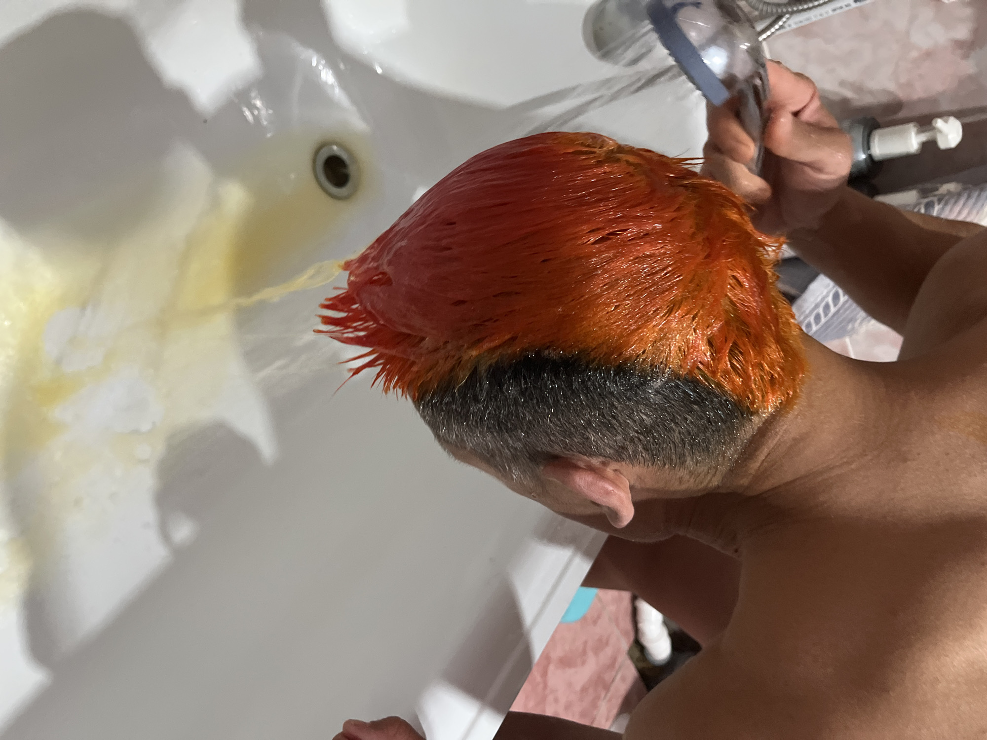 A person is rinsing orange hair dye from their hair in a white porcelain sink or bathtub. The drain water appears yellow. The lower portion of the person's hair is dark. The person is holding a shower head and leaning into the basin. Part of their torso and the side of their head are visible. The setting appears to be an interior bathroom, likely a private residence. There is no information in the image to determine if the photograph was taken in Chaikovsky, Russia.