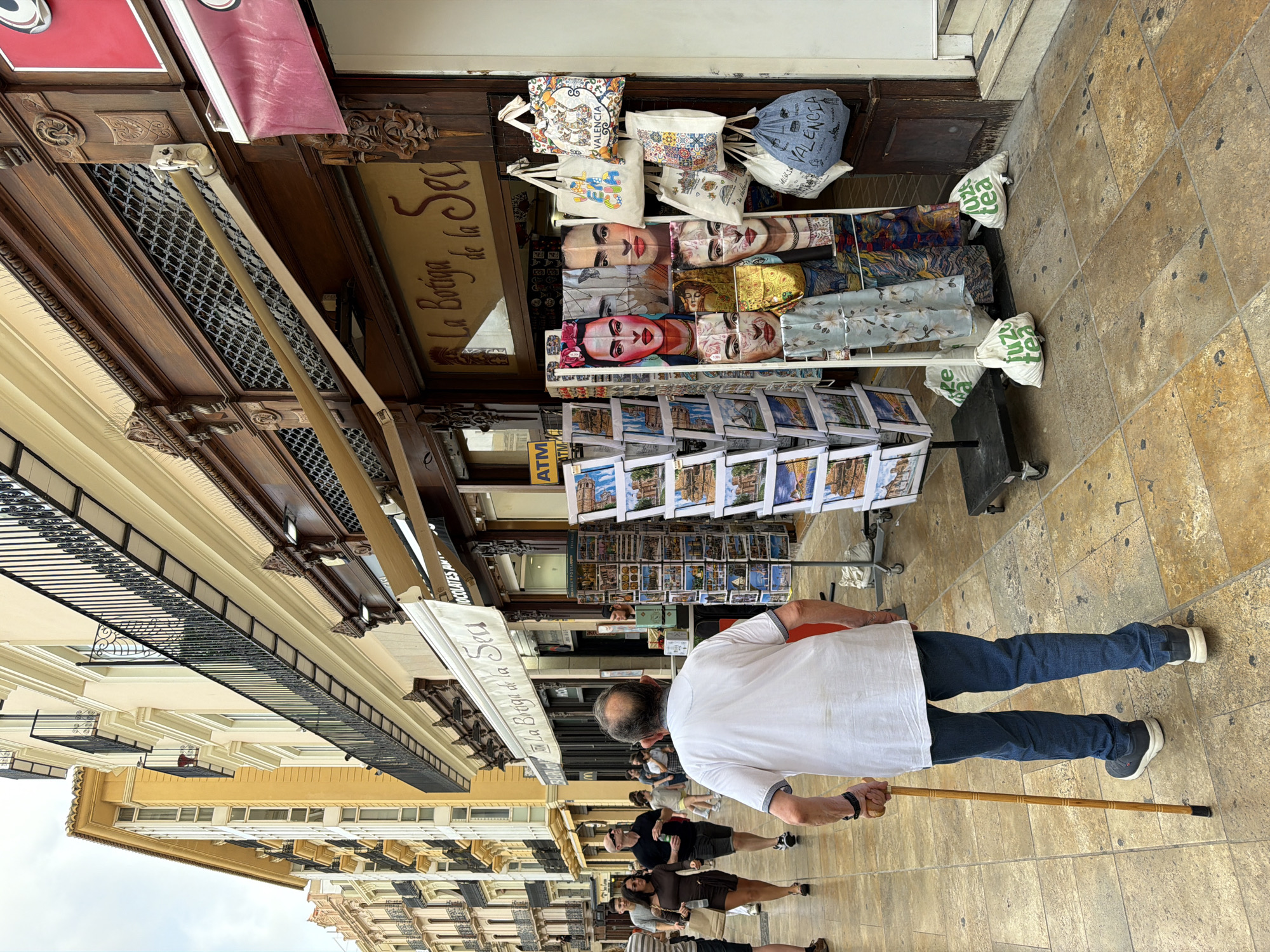 A street scene in a European city shows an elderly man with a cane walking towards the viewer on a tiled sidewalk. He is wearing a white short-sleeved shirt and blue jeans. To his right, a display rack holds postcards depicting scenic views, and next to it, fabric bags and scarves with artistic designs, including portraits of Frida Kahlo and images of Valencia, are hanging. The background features a building with balconies and shops, one of which has a sign reading "La Botiga de la Sec". People are visible walking in the background. The overall lighting suggests an overcast day.