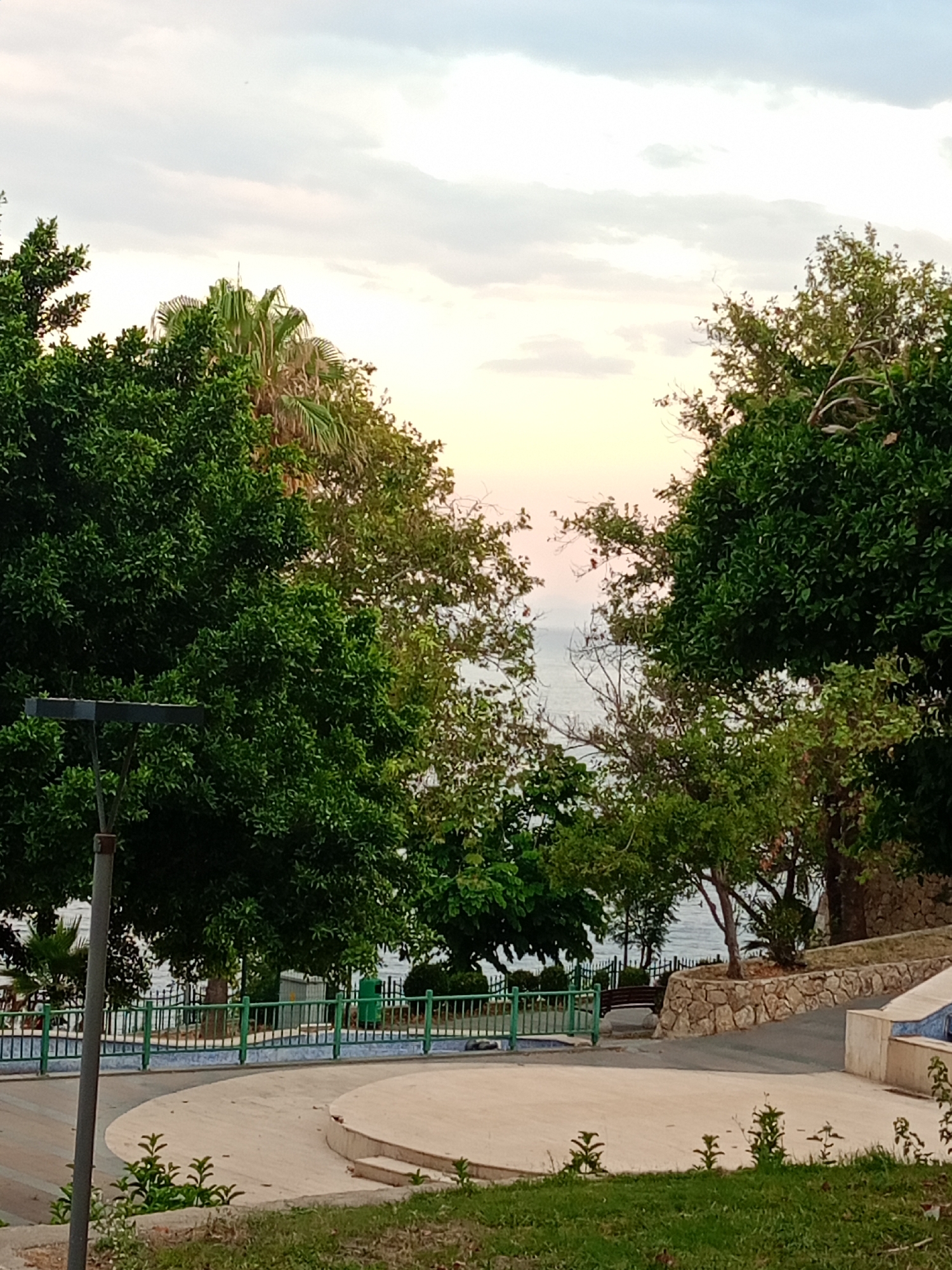 This image captures a serene park scene in Antalya, Turkey, overlooking the sea. In the foreground, a paved walkway with circular elements curves through lush green grass. To the left, a tall, dark lamppost stands prominently. Beyond the walkway, a vibrant green metal fence delineates the park's edge, behind which are more trees and glimpses of the sea. Several large trees, some with palm fronds visible, frame the view of the water. The sky is a soft gradient of pale yellow, pink, and blue, with scattered clouds, suggesting either dawn or dusk. The overall impression is one of a tranquil coastal public space.