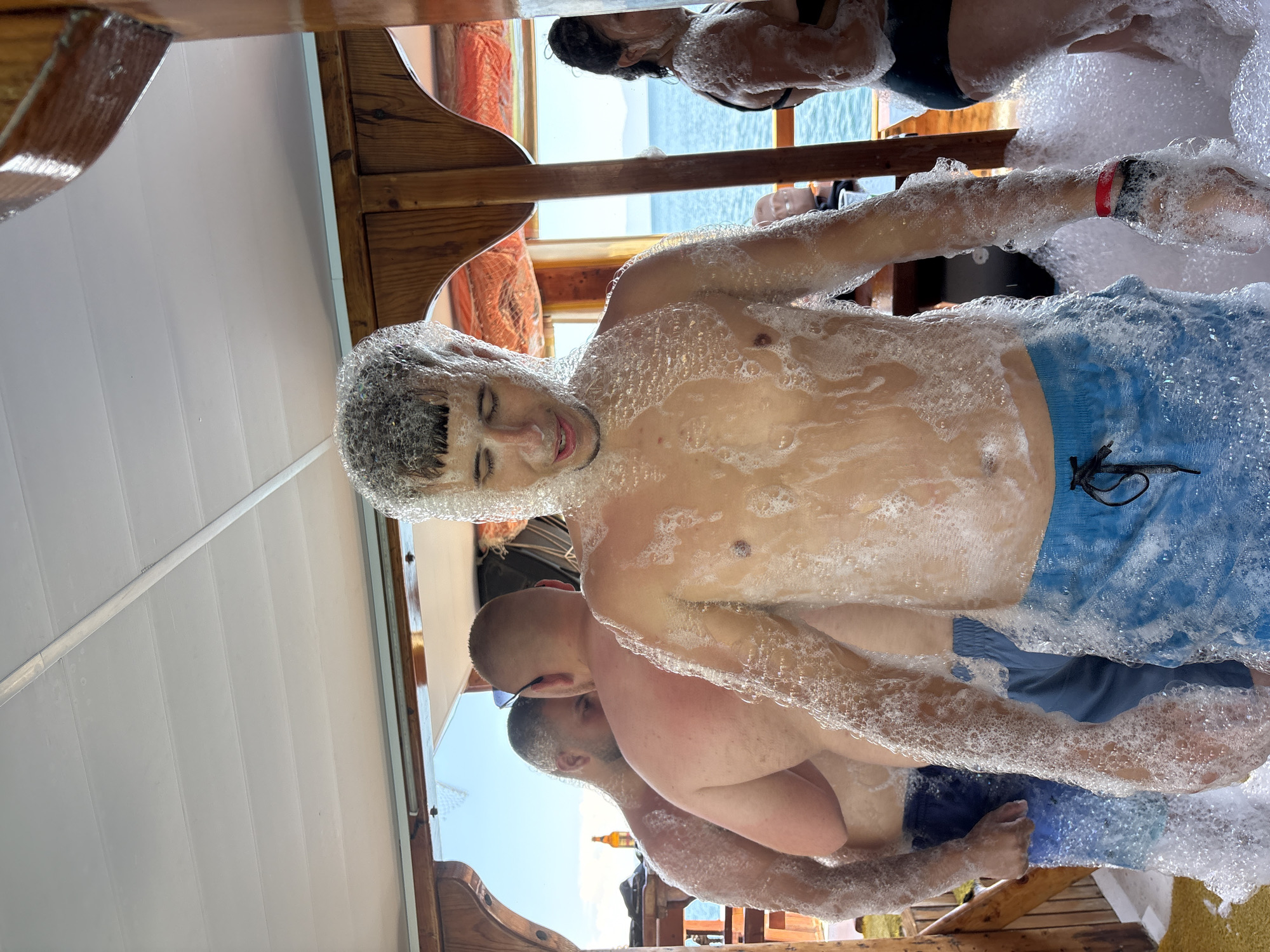 This image captures a vibrant foam party on a boat, likely off the coast of Menderes, Turkey. The primary subject is a young man, shirtless and wearing blue swim shorts, covered head-to-toe in white foam. He appears to be smiling with his eyes closed, suggesting enjoyment. His hair is also heavily coated in foam.
In the background, several other individuals are visible, similarly covered in foam. One person, partially obscured, wears a black top and is also covered in suds. Another man with a shaved head and sunglasses rests his arm on a wooden railing, also covered in foam, with another person behind him. A red wristband is visible on the arm of one person. The boat features wooden railings and structures, indicating a classic boat or gulet design. The ceiling above is white with horizontal panels.
Beyond the boat, the bright blue sea stretches out under a clear, sunny sky, with distant land or mountains visible on the horizon, confirming a coastal setting. The overall atmosphere suggests a lively and fun daytime event. There is no visible text in the image. A yellow bottle can be faintly seen in the reflection of the boat's polished surface in one of the cropped images.