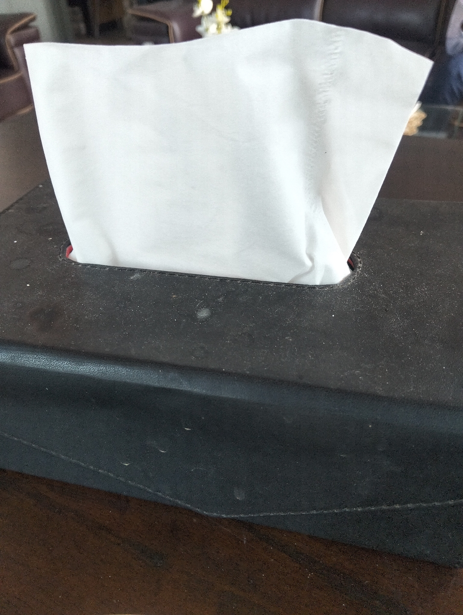 A black tissue box with white tissues protruding from the top sits on a dark wooden surface. The tissue box is rectangular and appears to be made of faux leather. There are small specks of dust visible on the surface of the tissue box. The background is blurred, but suggests an indoor setting with furniture. The image is taken from a slightly elevated perspective, looking down at the tissue box. There is no indication of any specific location in the city of Julicotmeurak, Indonesia.