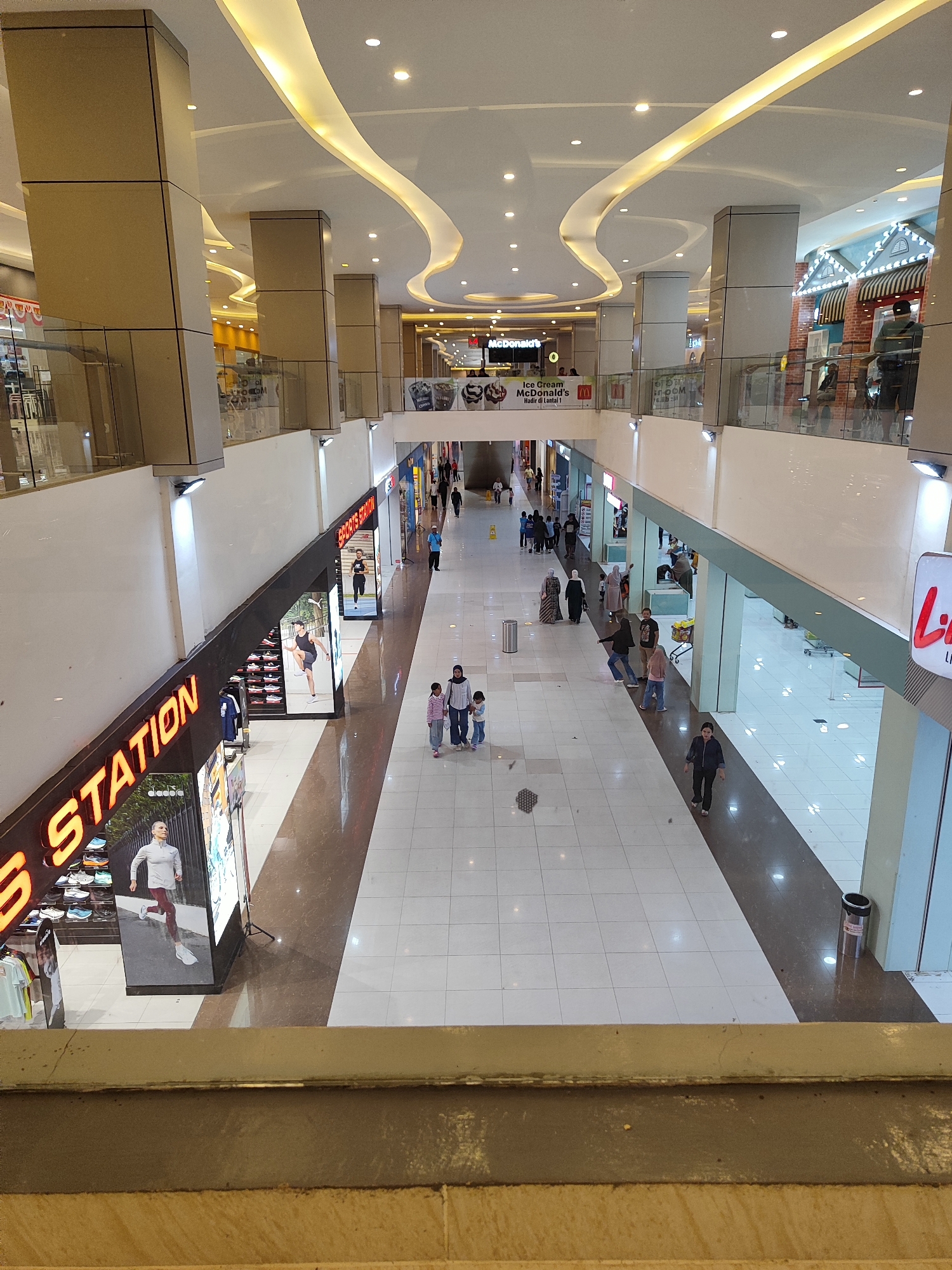 The image shows an interior view of a modern shopping mall, likely located in Sempusari, Indonesia, as suggested by the visible signage. The scene is captured from an elevated vantage point, looking down a wide, well-lit corridor.

In the foreground, to the left, is a store with prominent orange neon lettering that reads "STATION". Part of the store's display is visible, featuring athletic shoes and apparel. A large advertising display shows a woman in athletic wear.

The mall's architecture is characterized by large, square pillars clad in a light-colored material, supporting a high ceiling with a distinctive, undulating wave design illuminated by warm yellow LED lighting. Numerous recessed circular lights are also present in the ceiling. The floor is a combination of polished white tiles and dark brown marble-effect tiles, creating a path through the central area.

Several individuals are visible in the mall. A group of three people, two adults and a child, are walking together in the middle distance on the white tiled area. Further down the corridor, more people are seen browsing or walking. On the right side of the image, a store with red lettering, partially obscured, is visible. There are also store fronts and displays along both sides of the corridor.

In the upper background, a McDonald's restaurant is advertised with its signature golden arches logo and signage that includes Indonesian text. Other commercial establishments are also identifiable in the distance. Trash receptacles are present along the corridor, one is visible on the right side. The overall environment suggests a busy, well-maintained commercial space.